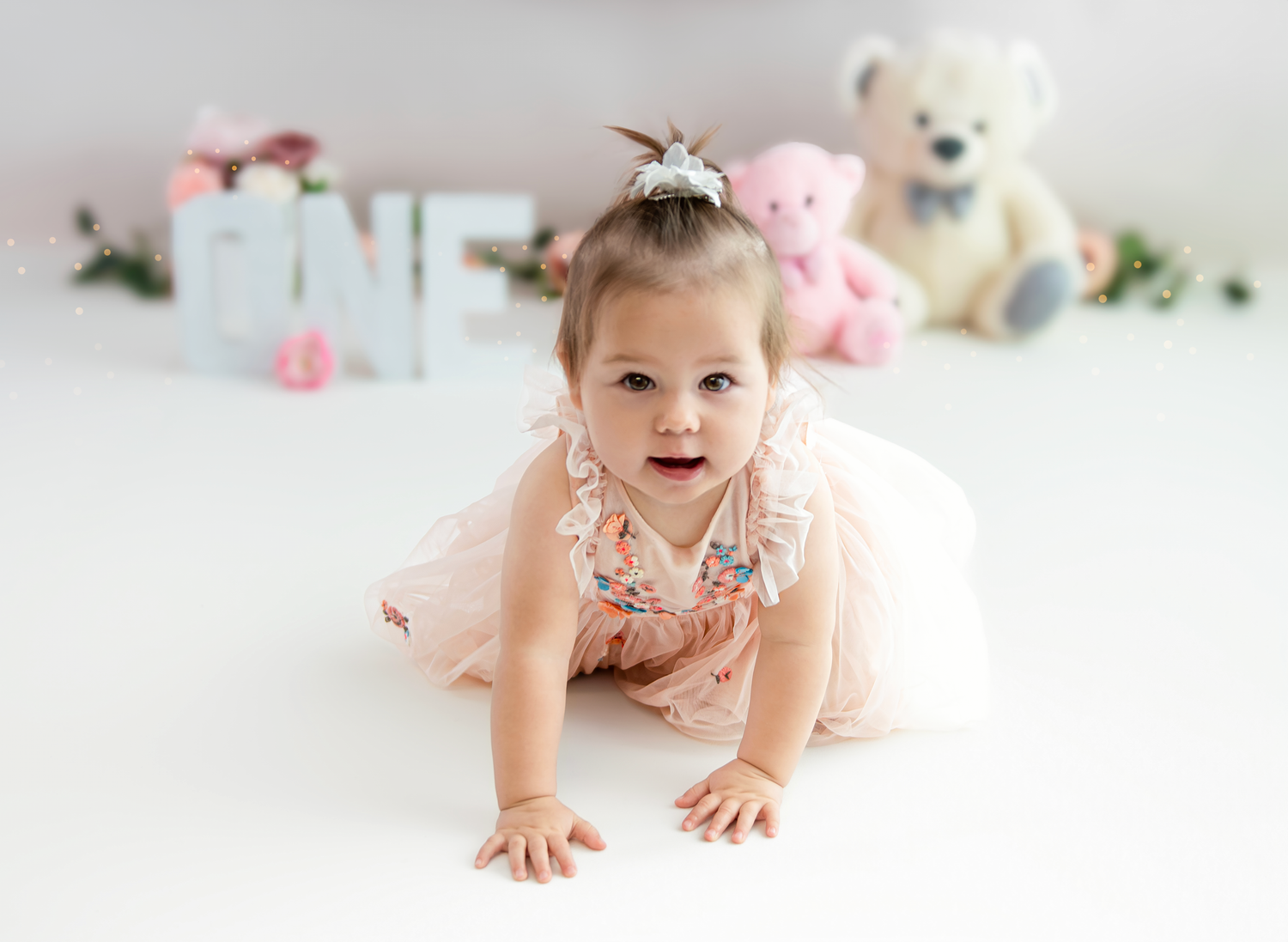 A small child in a floral dress crawls on a white floor with stuffed animals and decorations in the background.