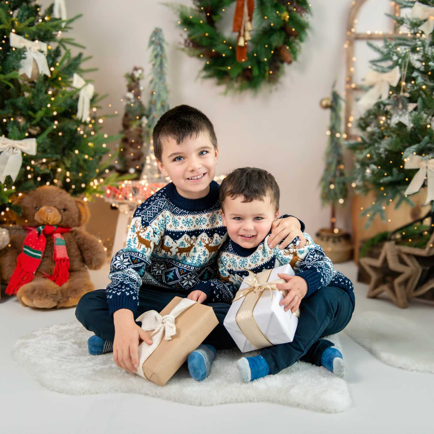 Two children in matching blue sweaters pose together on a white floor with Christmas decorations in the background.