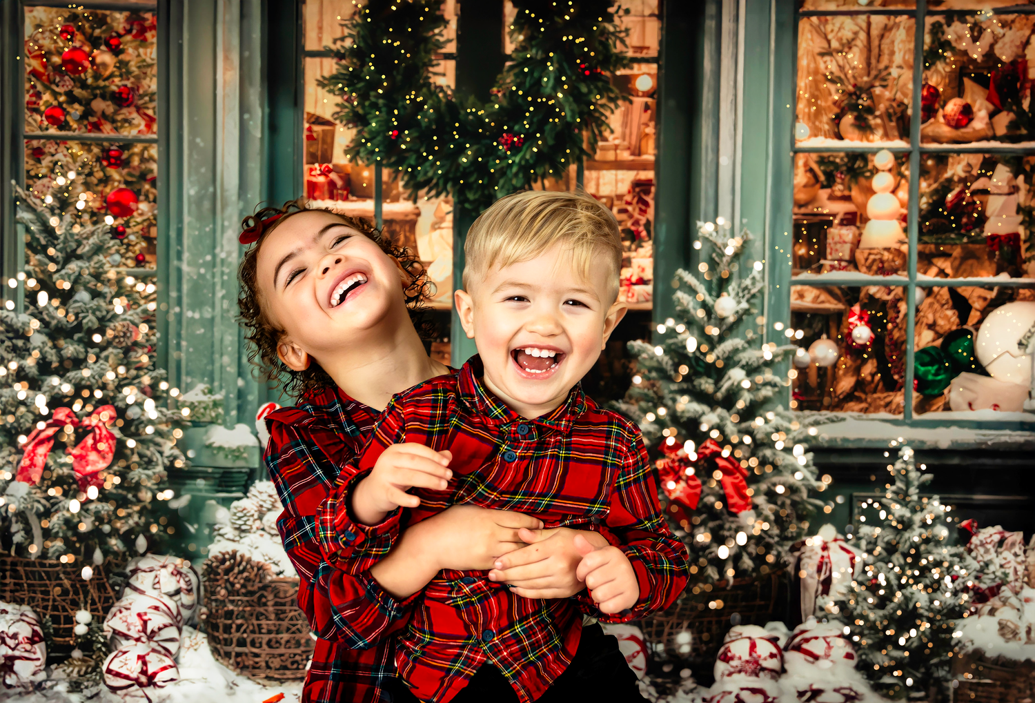 Two people wearing matching red plaid outfits laugh joyfully in front of a festive Christmas window display with wreaths.
