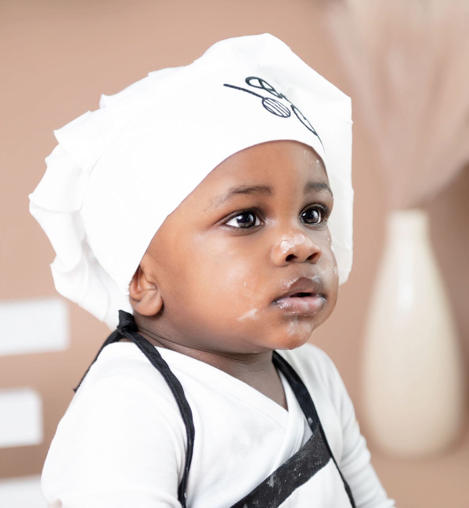 Baby in white chef's hat and black apron smiles while cooking in bright kitchen setting.