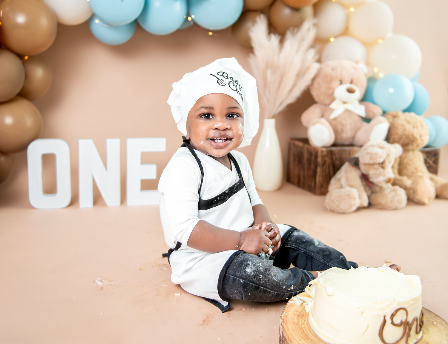 A baby in a chef outfit sits near decorative 'ONE' letters and balloon garlands with stuffed animals in the background.