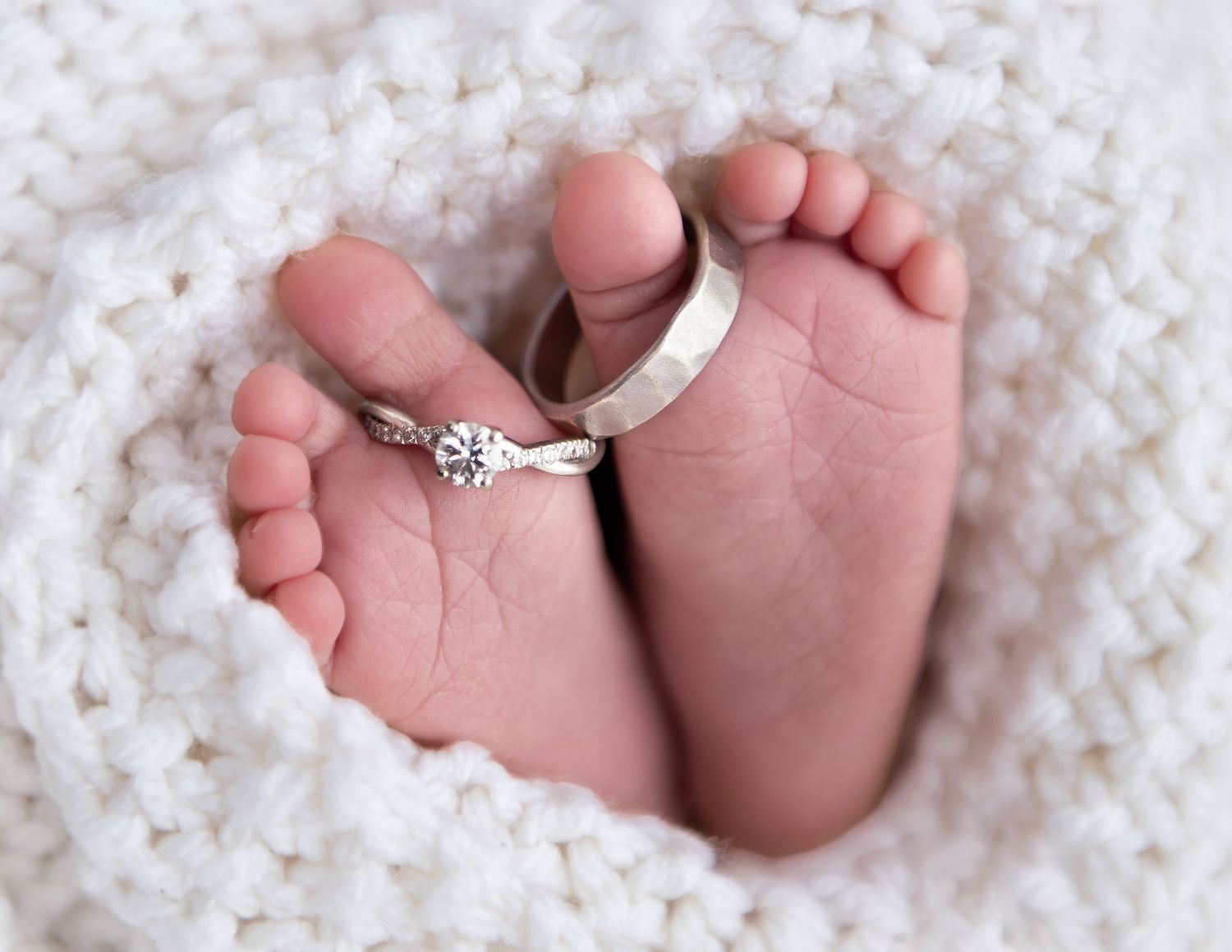 Newborn baby feet wrapped in white knit blanket with diamond engagement ring and silver wedding band.
