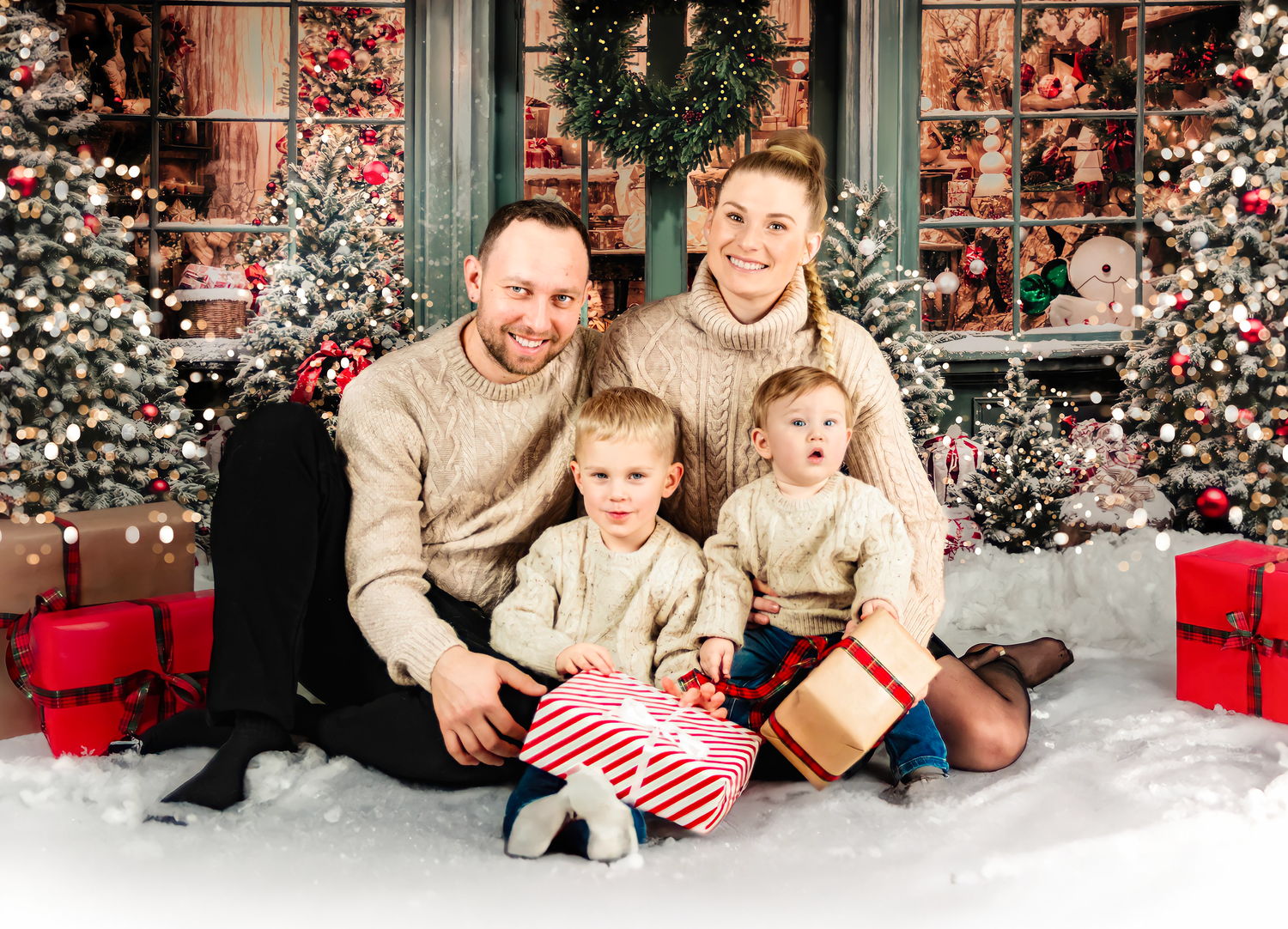 A cozy family holiday portrait with matching cream sweaters sitting by decorated Christmas trees and red gift boxes.