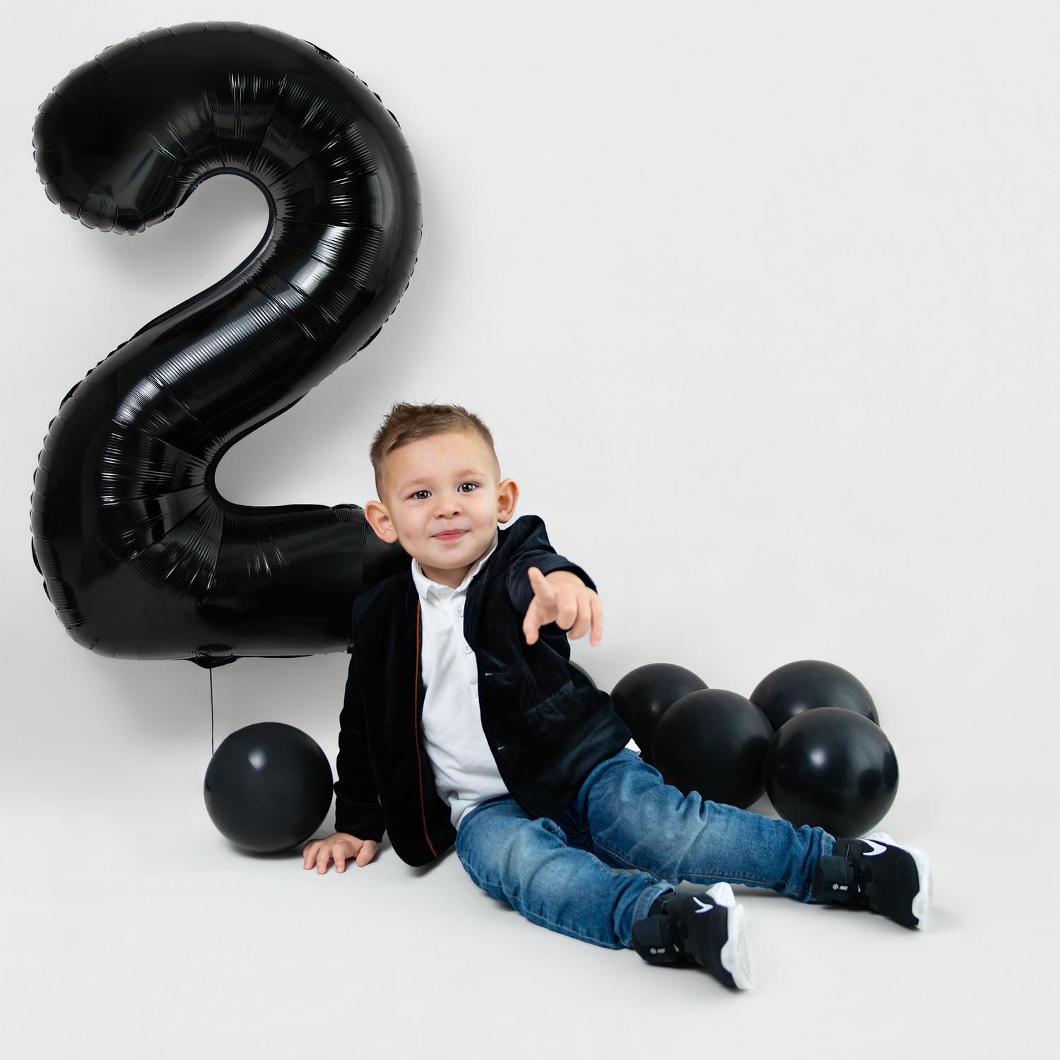 A child poses next to a large black number 2 balloon with black party balloons on a white backdrop.