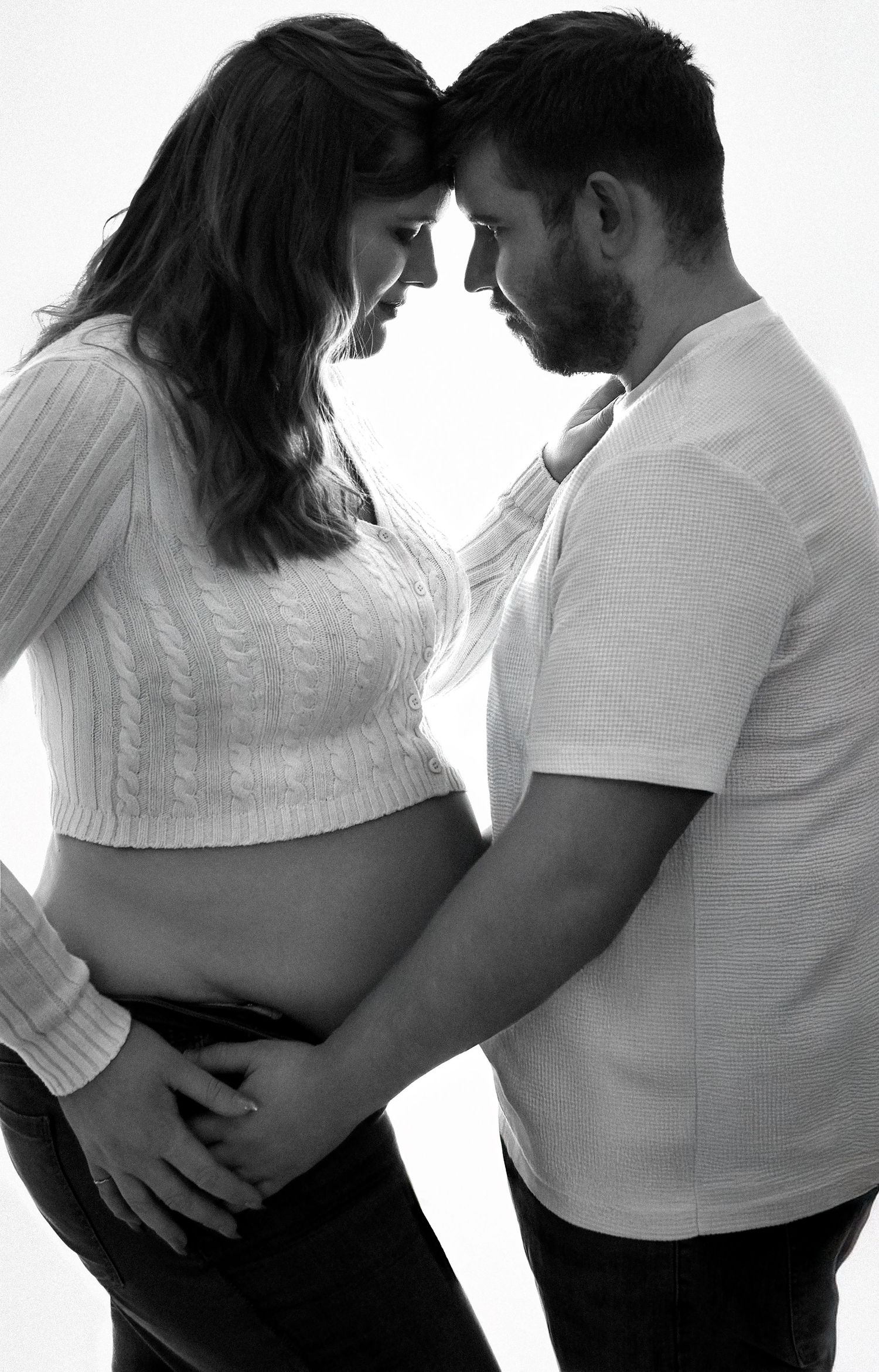 Black and white maternity photo of pregnant couple tenderly touching foreheads while cradling baby bump.