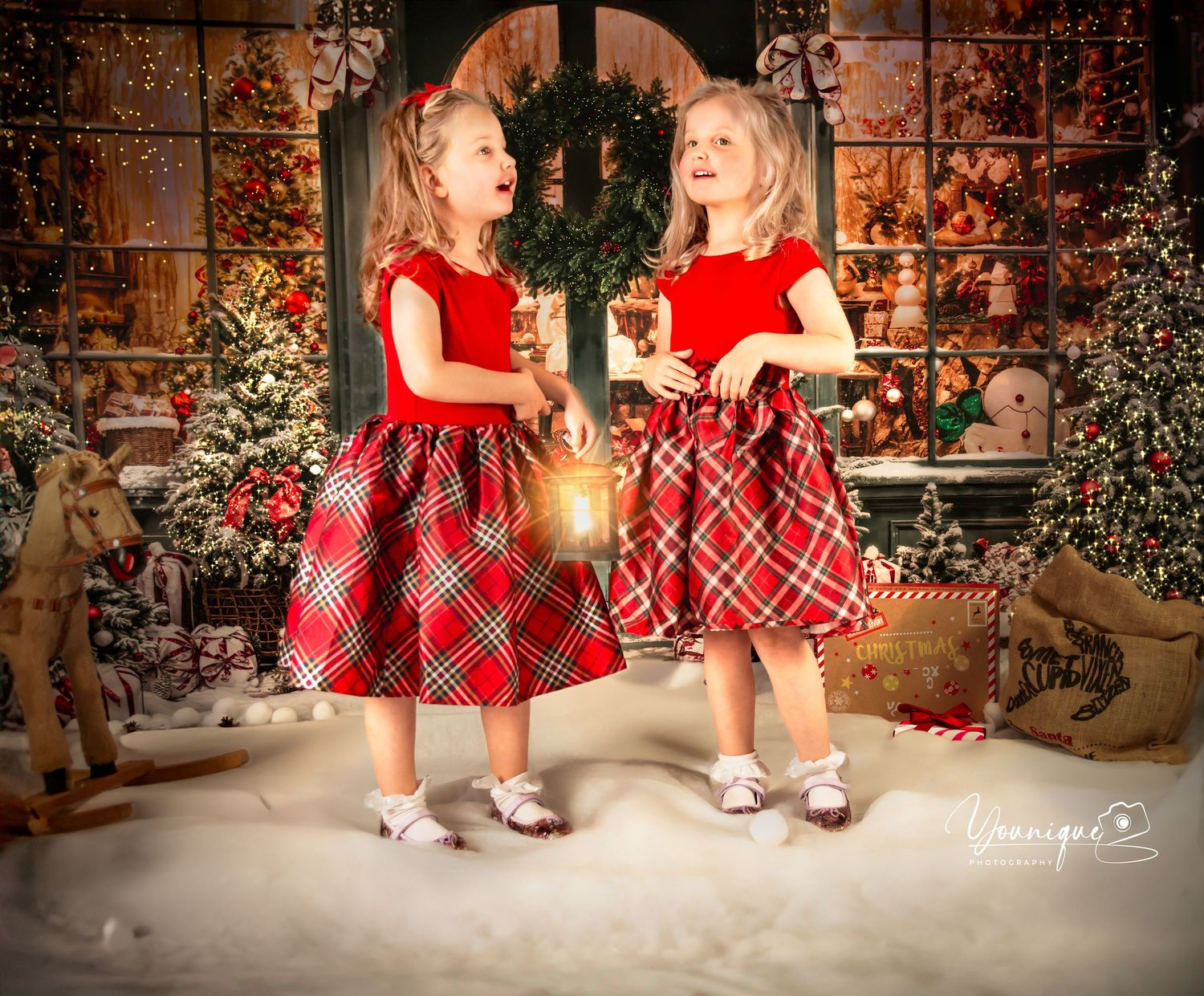 Two children in matching red plaid dresses pose in a festive Christmas studio setting with snow and decorations.