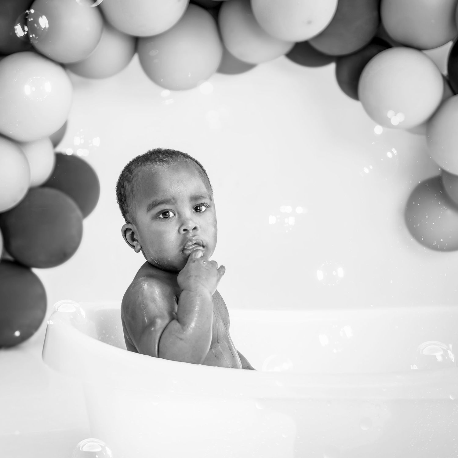Black and white photo of a baby sitting in a small tub surrounded by balloons and bubbles.