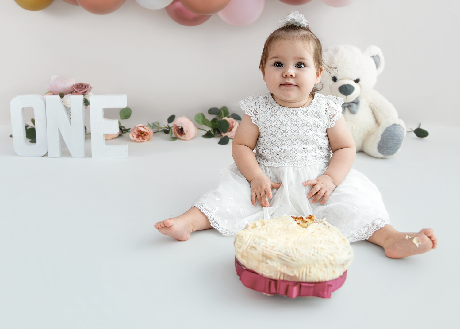 A baby in a white dress sits on the floor with stuffed animals and toys during a cake smash photo session.