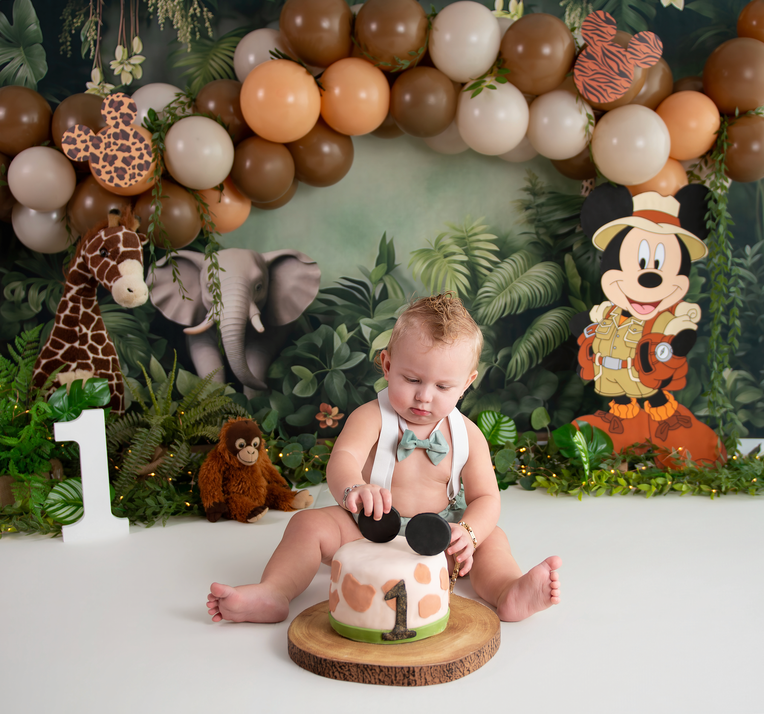 A baby's first birthday woodland theme photo session with Mickey Mouse plush toy and balloon decorations in earth tones.