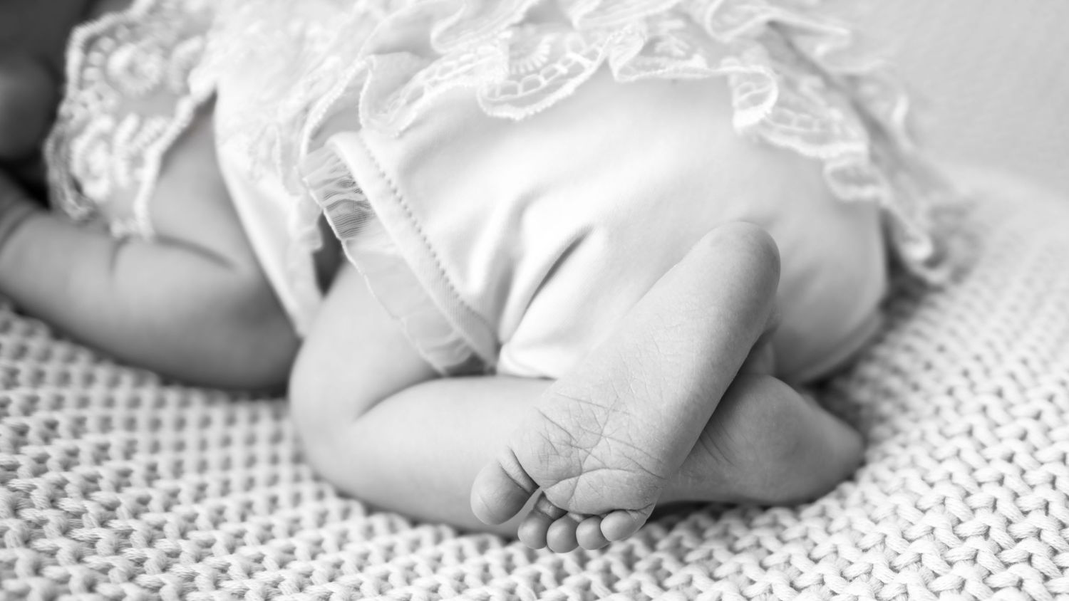 Black and white photo of a sleeping newborn baby's tiny feet and hands in a lace outfit on a knit blanket.