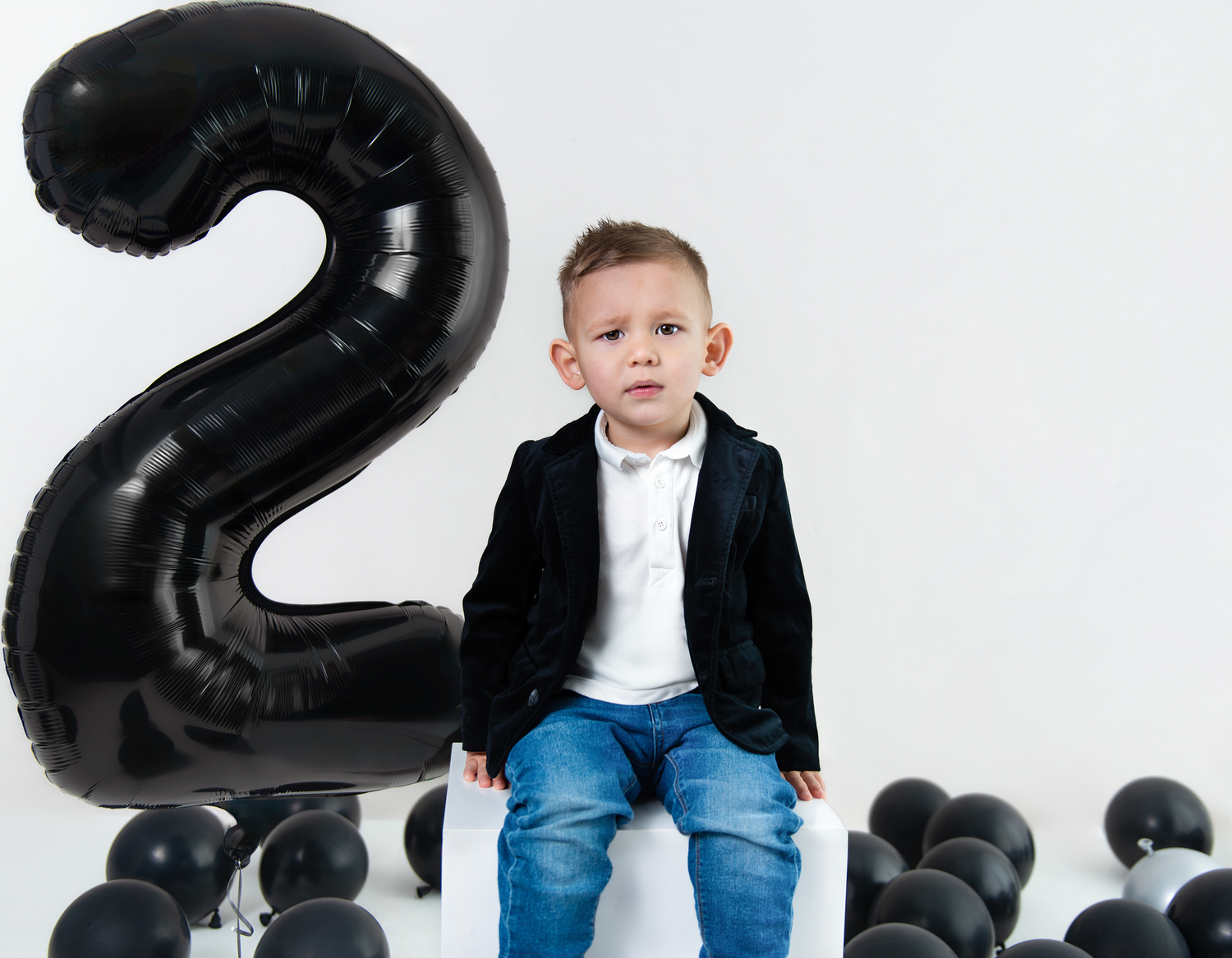 A child in a black blazer and jeans sits next to a large black number 2 balloon with black balloons on the floor.