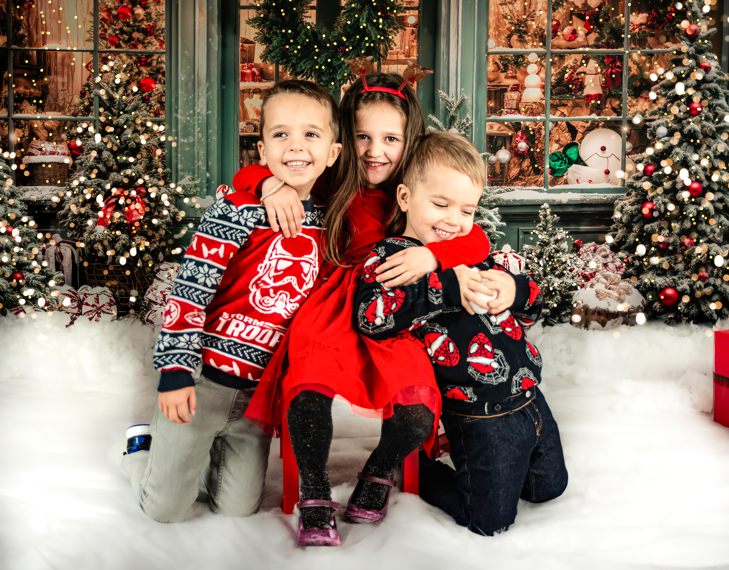 Three children wearing festive winter sweaters pose together in front of Christmas decorations and wreaths.