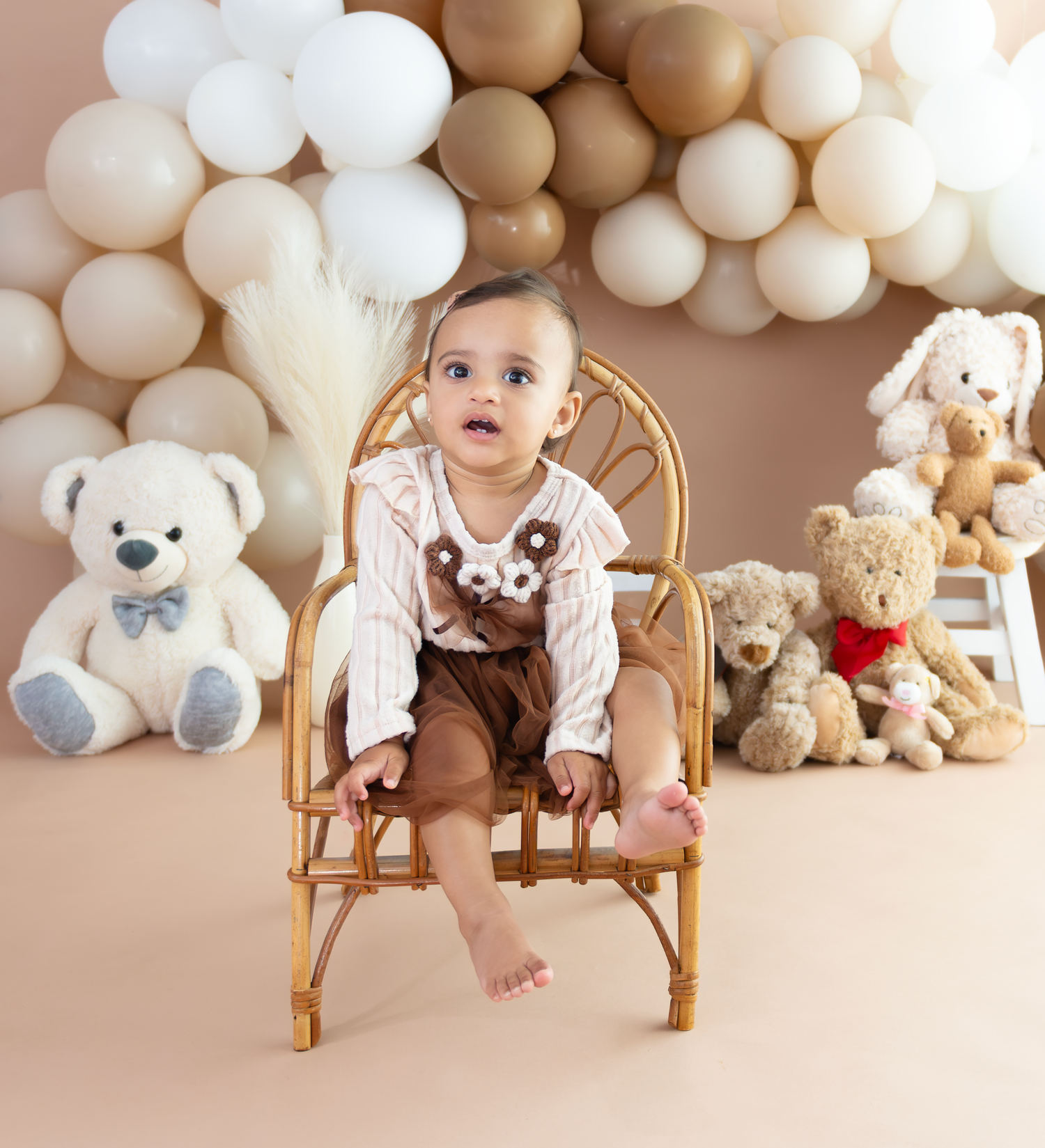 A festive first birthday setup with teddy bears, wooden rocking chairs, and white balloon decorations against a neutral backdrop.