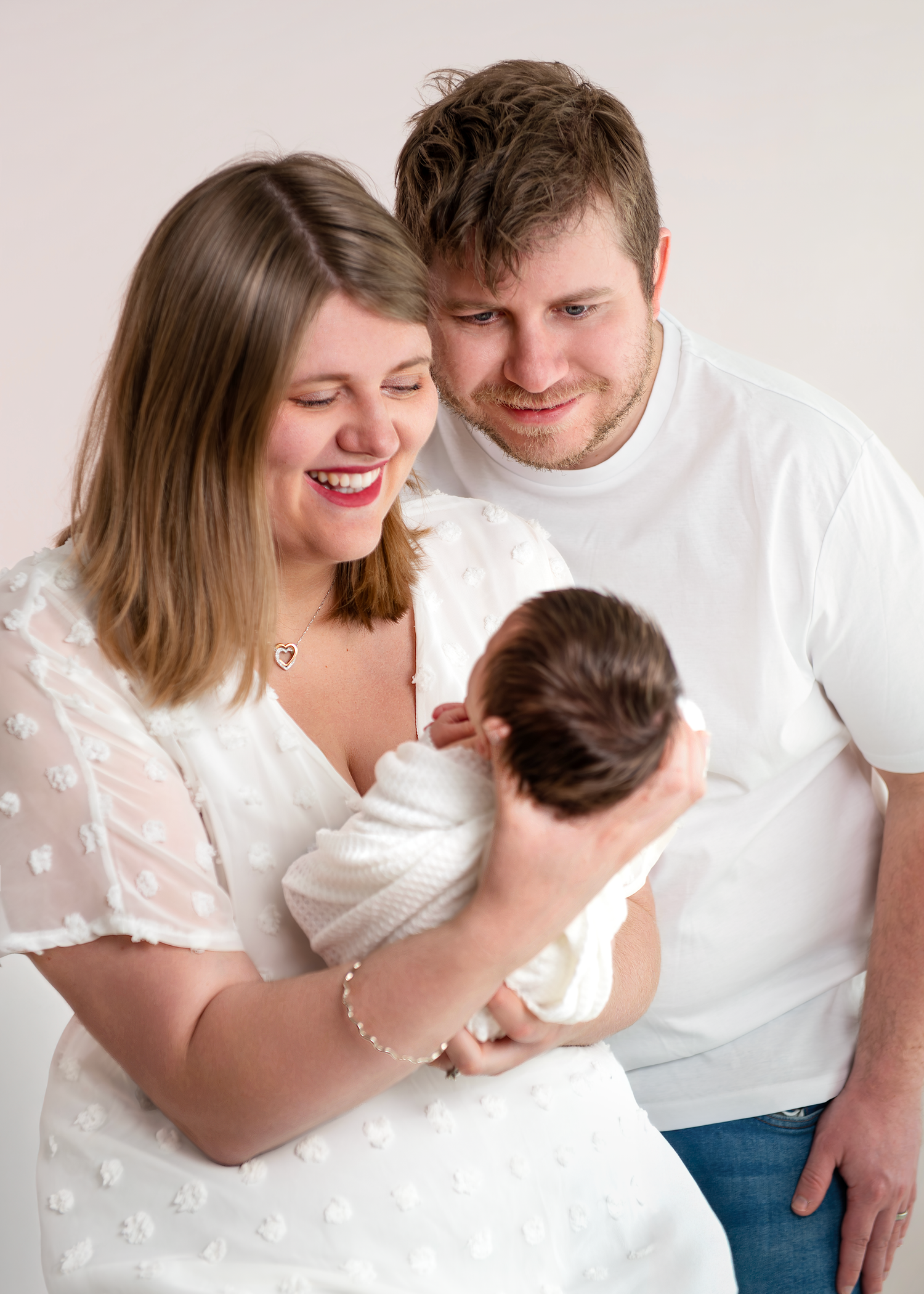 Happy parents in white outfits lovingly gaze at their newborn baby during a professional studio portrait session.