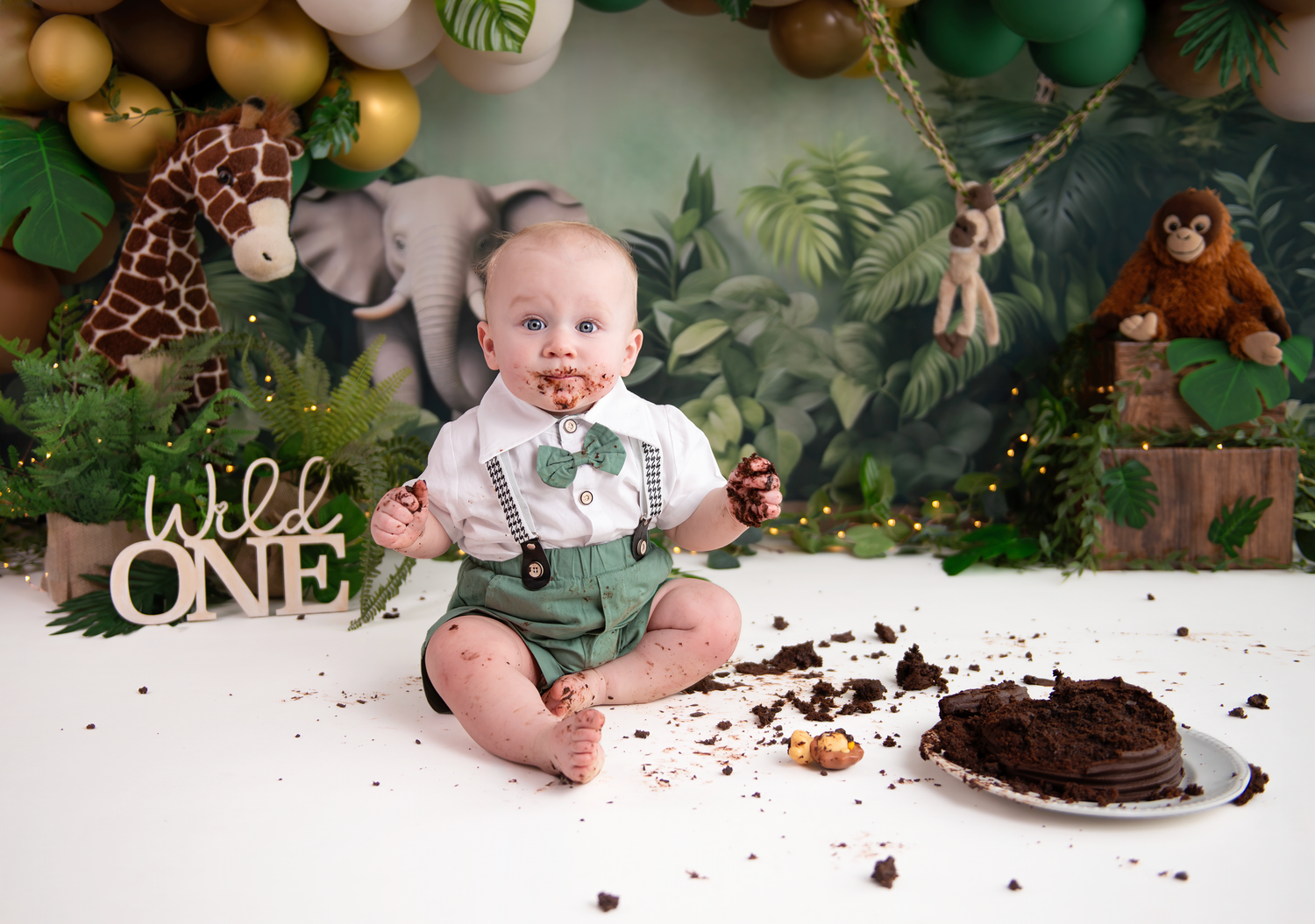 Baby boy in green suspenders smashes chocolate cake at Wild One jungle themed first birthday photo shoot.