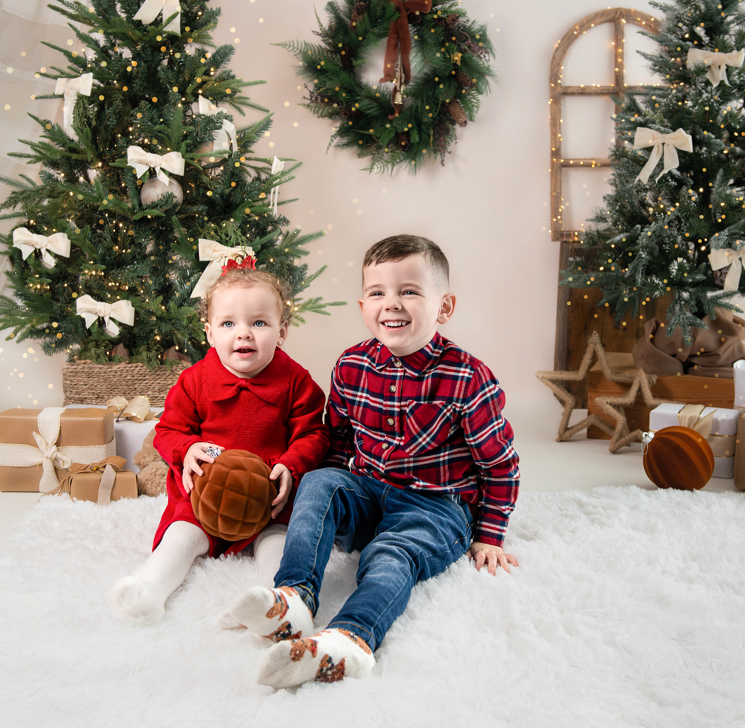 Children in red and plaid clothing play and laugh together in a winter holiday setting with wreaths and decorative snow.