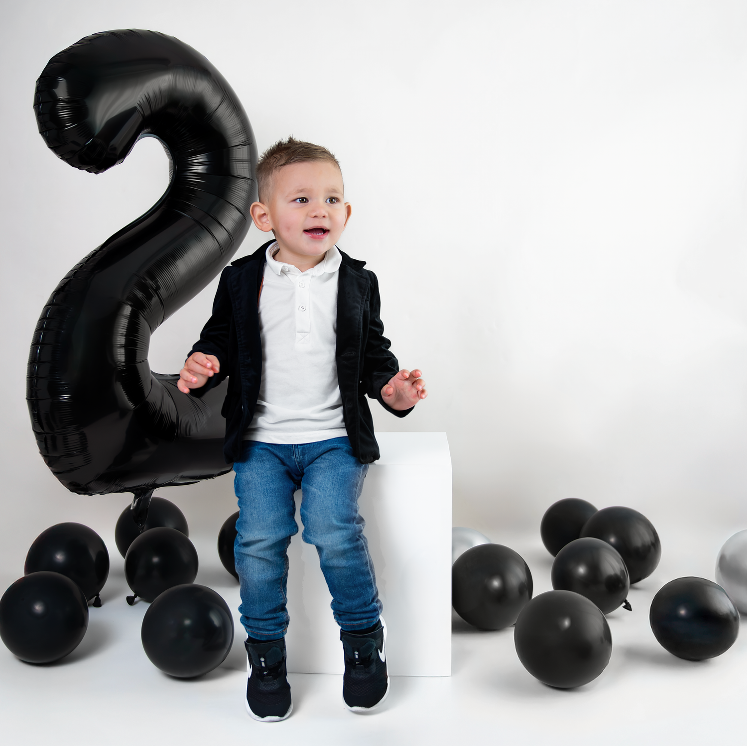 A sequence of black balloon numbers and a toddler in jeans and jacket standing against a white backdrop for a photo shoot.