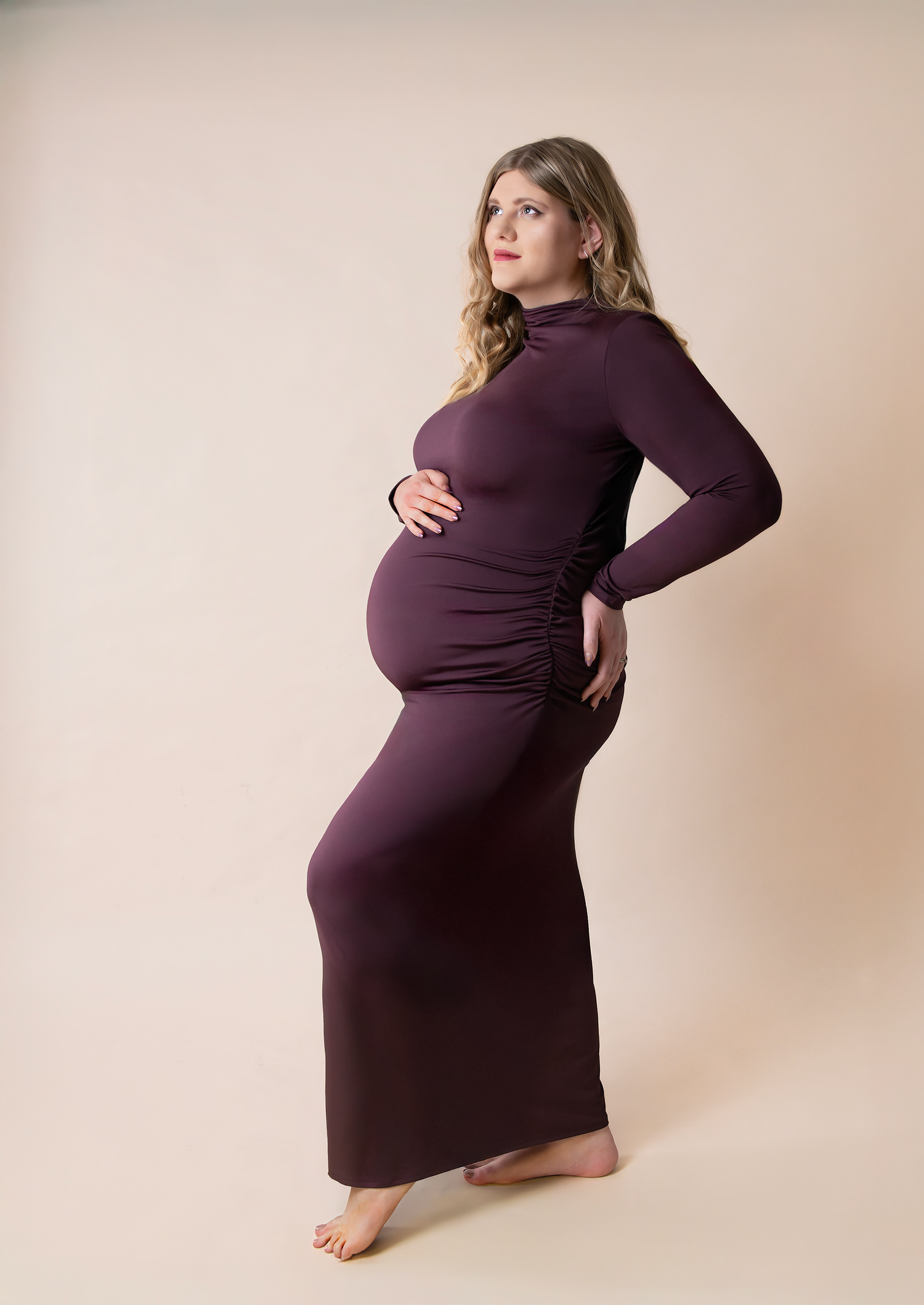 Pregnant woman in a dark plum long-sleeve maxi dress posing for a maternity photoshoot on a beige background.