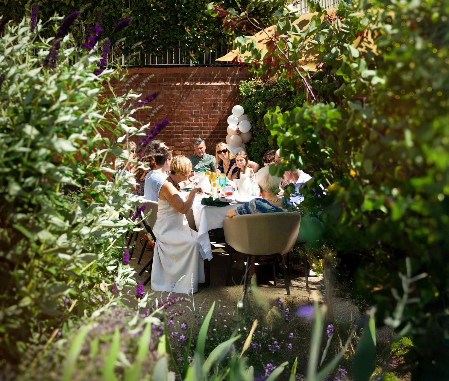 Garden party scene with people seated around a table surrounded by greenery.
