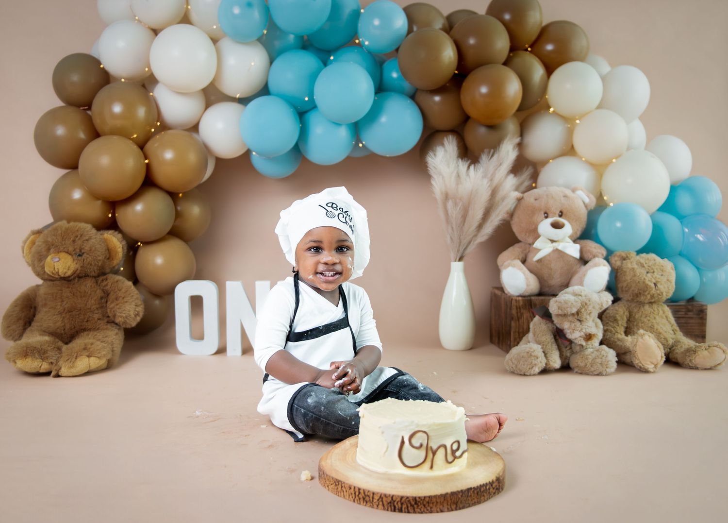 A festive first birthday cake smash setup with blue and brown balloon garlands and teddy bear decorations against beige backdrop.