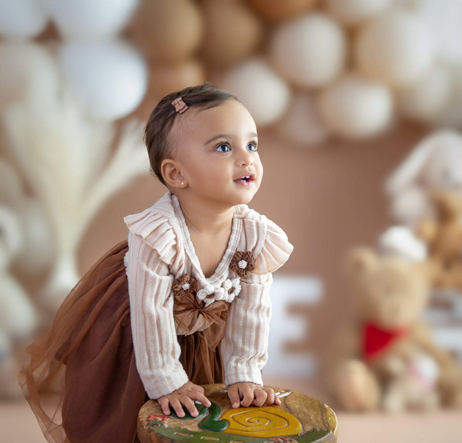 A child in a white dress with a brown bow smiles and looks up while sitting against a soft neutral background.