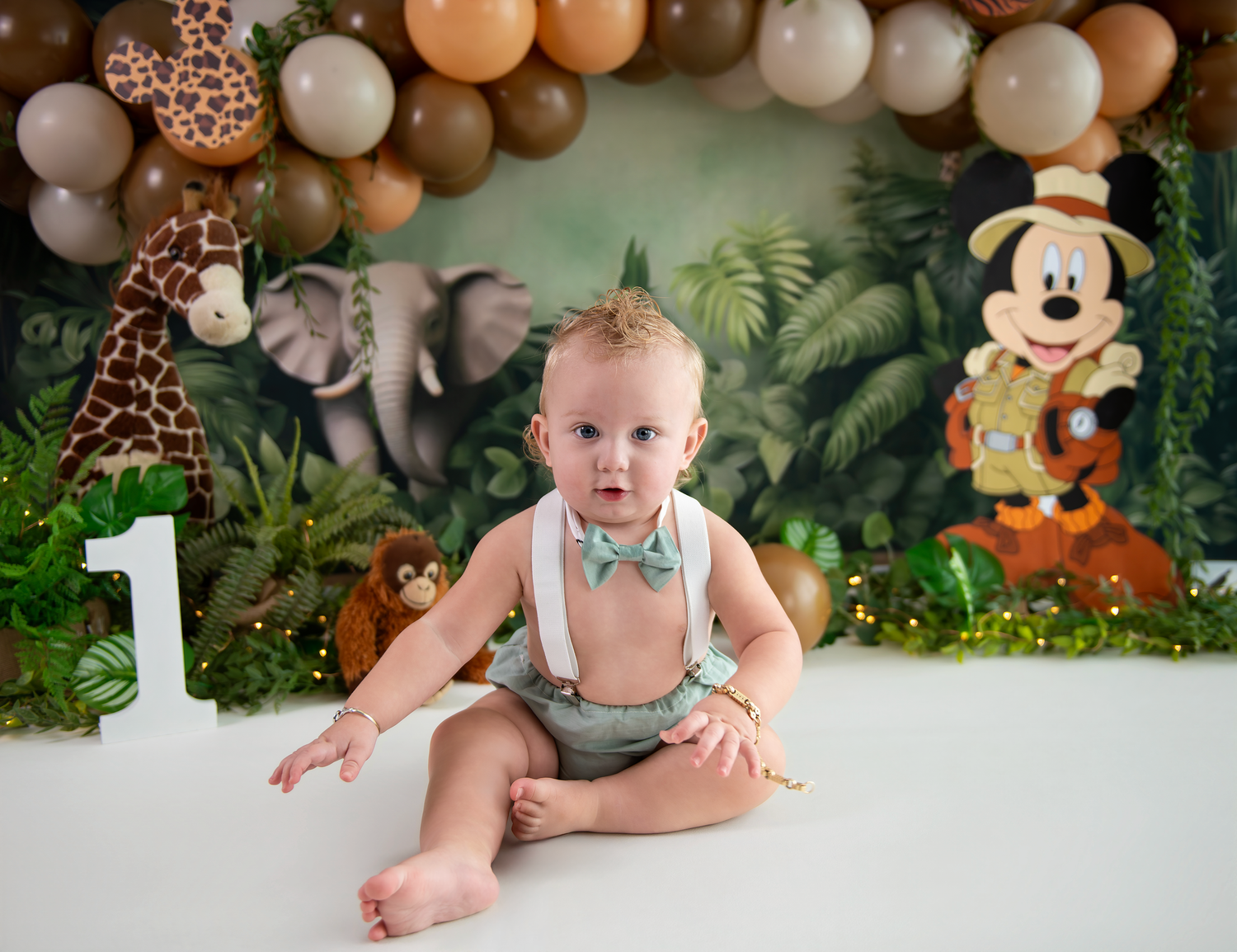 Safari themed first birthday party setup with balloon arch, jungle animals and Mickey Mouse decorations against a tropical backdrop.