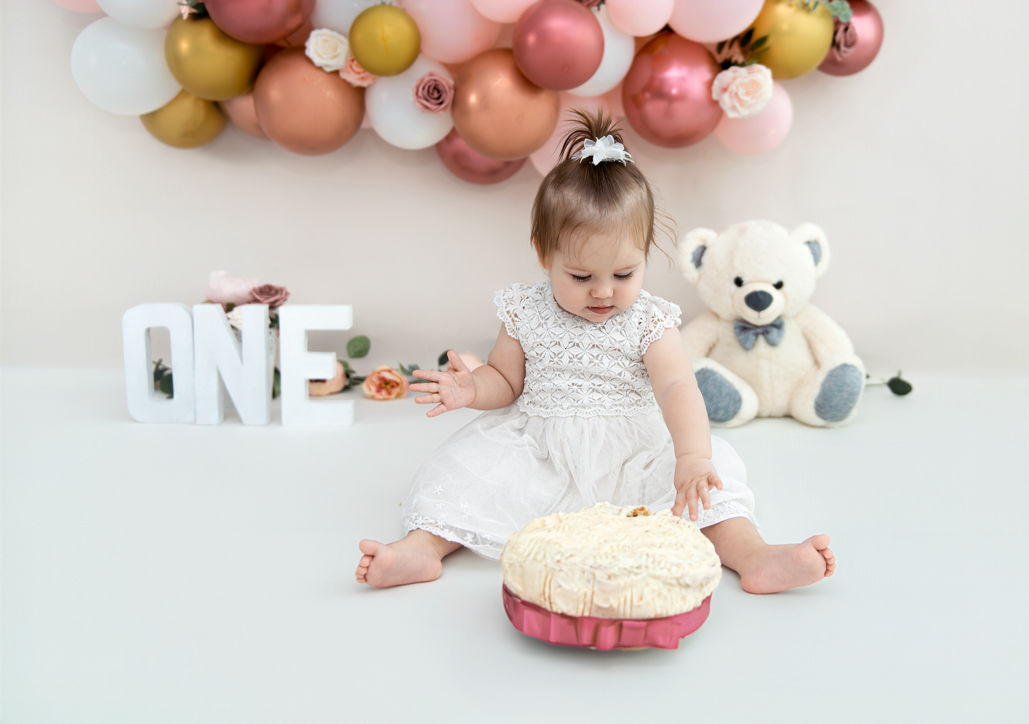 A baby in white lace dress sits on a white blanket with stuffed animals and a pink birthday cake for a photo session.