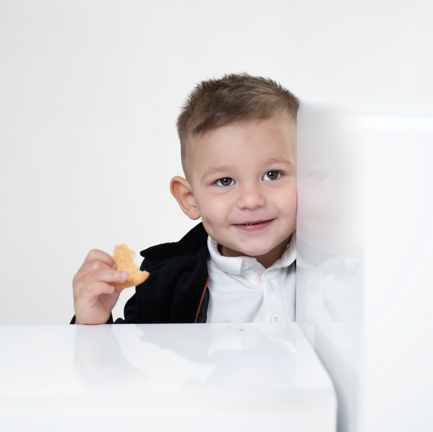 Young child in formal attire with white bowtie smiling while holding a small snack against white background.