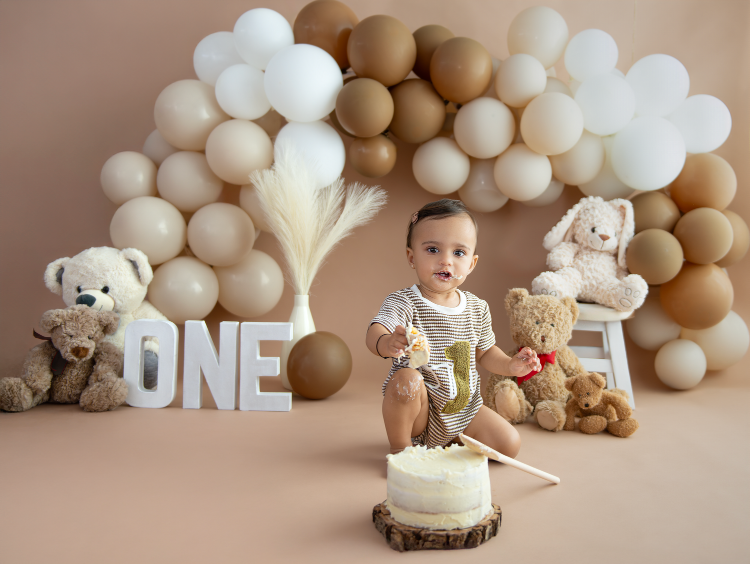 A baby's first birthday photoshoot setup with neutral beige balloons, teddy bears, and a rustic wood slice cake stand.