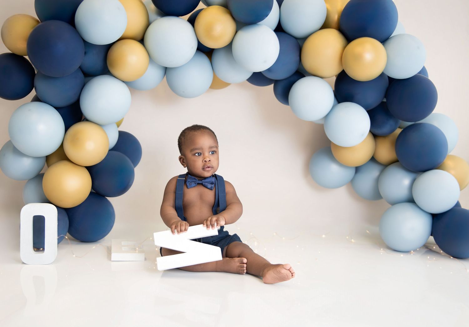 Baby boy in bow tie sits before blue and gold balloon arch for first birthday cake smash photo.
