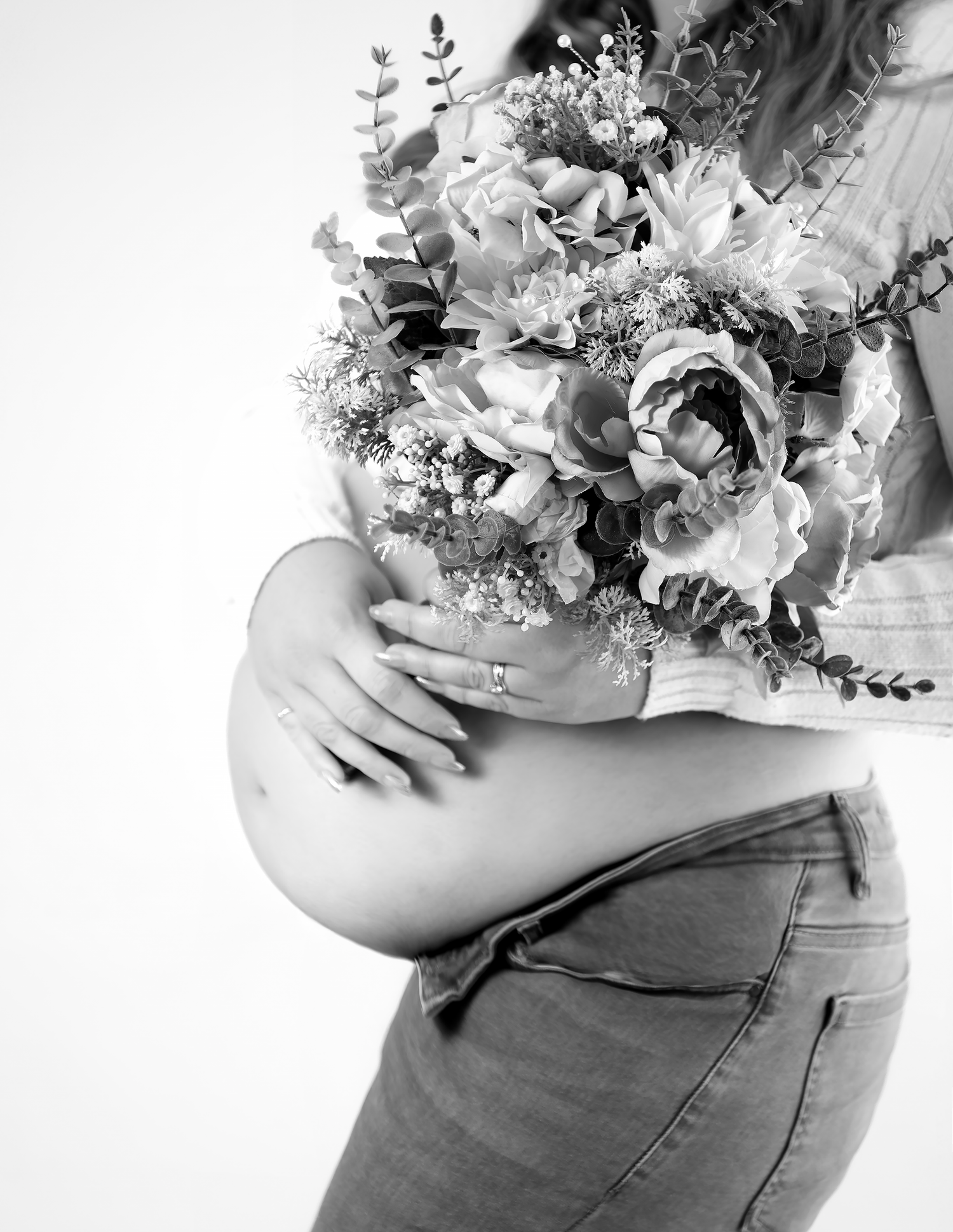Black and white maternity photo of pregnant woman holding flower bouquet, wearing jeans, showing baby bump.