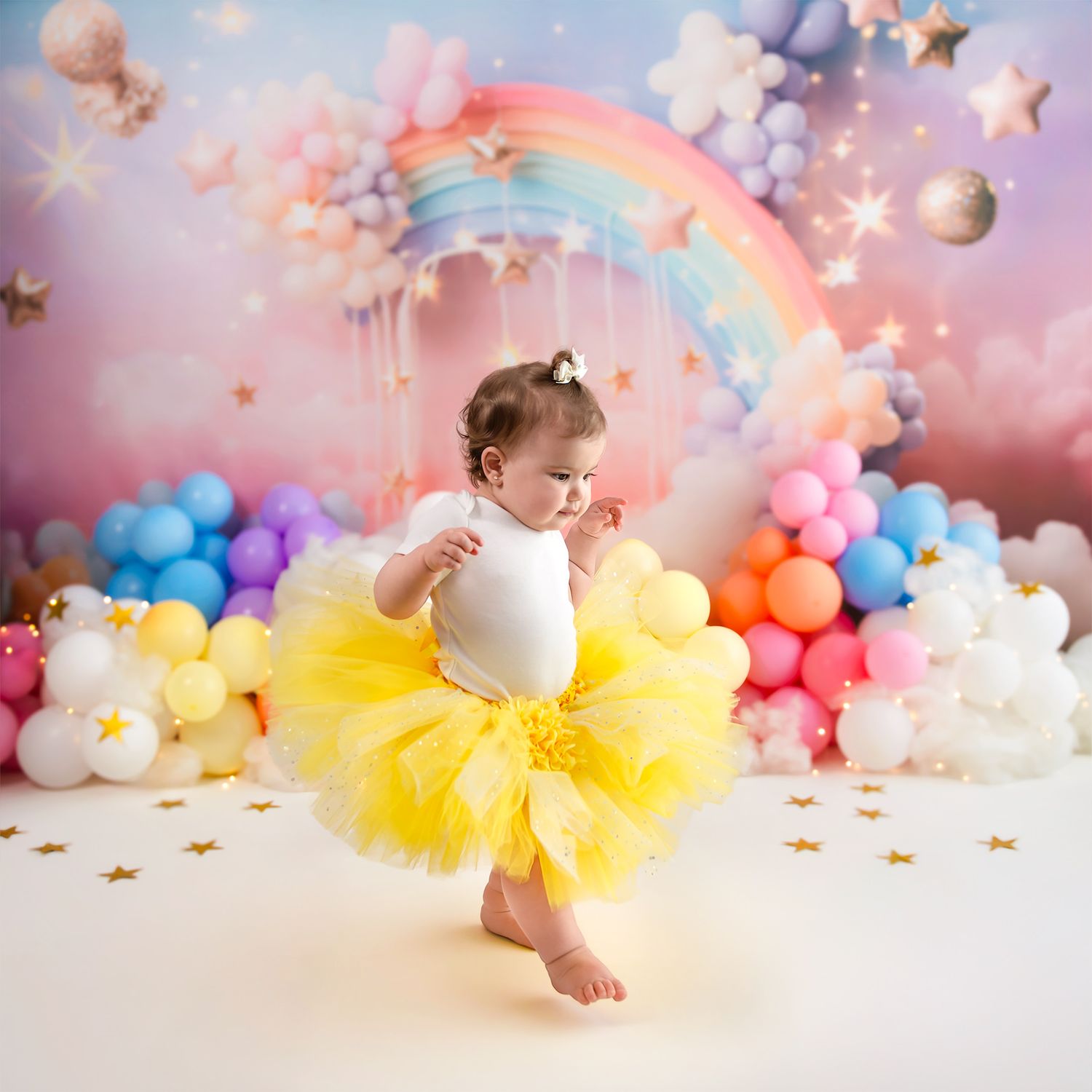 Baby girl in yellow tutu dress stands before colorful balloon arch with rainbow backdrop at first birthday cake smash.