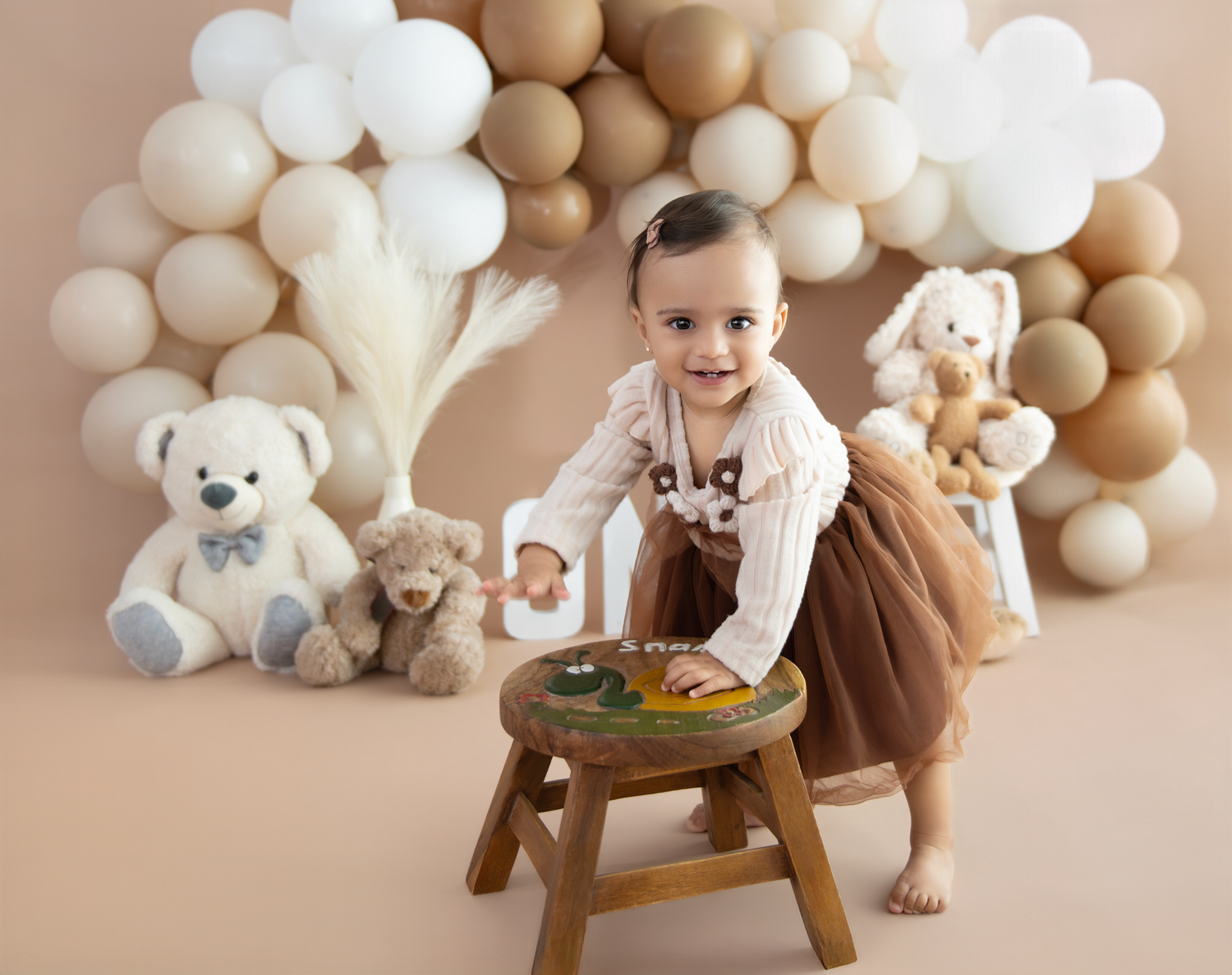 Baby in brown outfit sits on wooden chair with teddy bears and neutral balloon arch backdrop for first birthday photos.