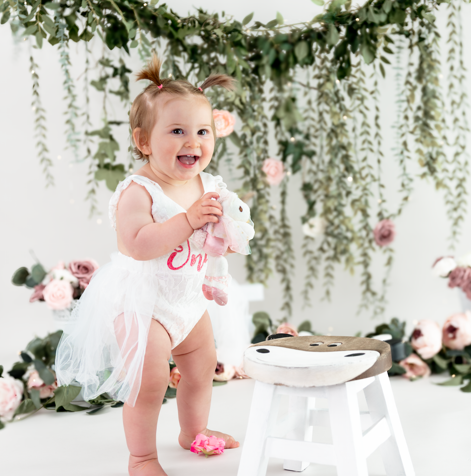 A child in a white dress sits at an elegant table setting with hanging floral decor and greenery above.