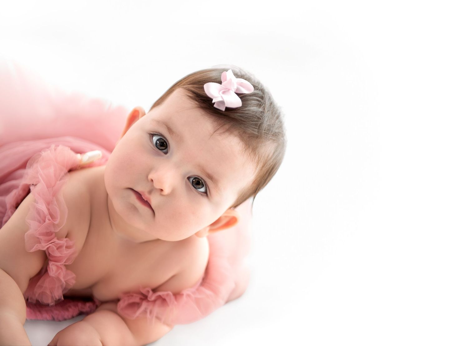 Adorable baby girl in pink tutu dress with flower headband lying on white background.