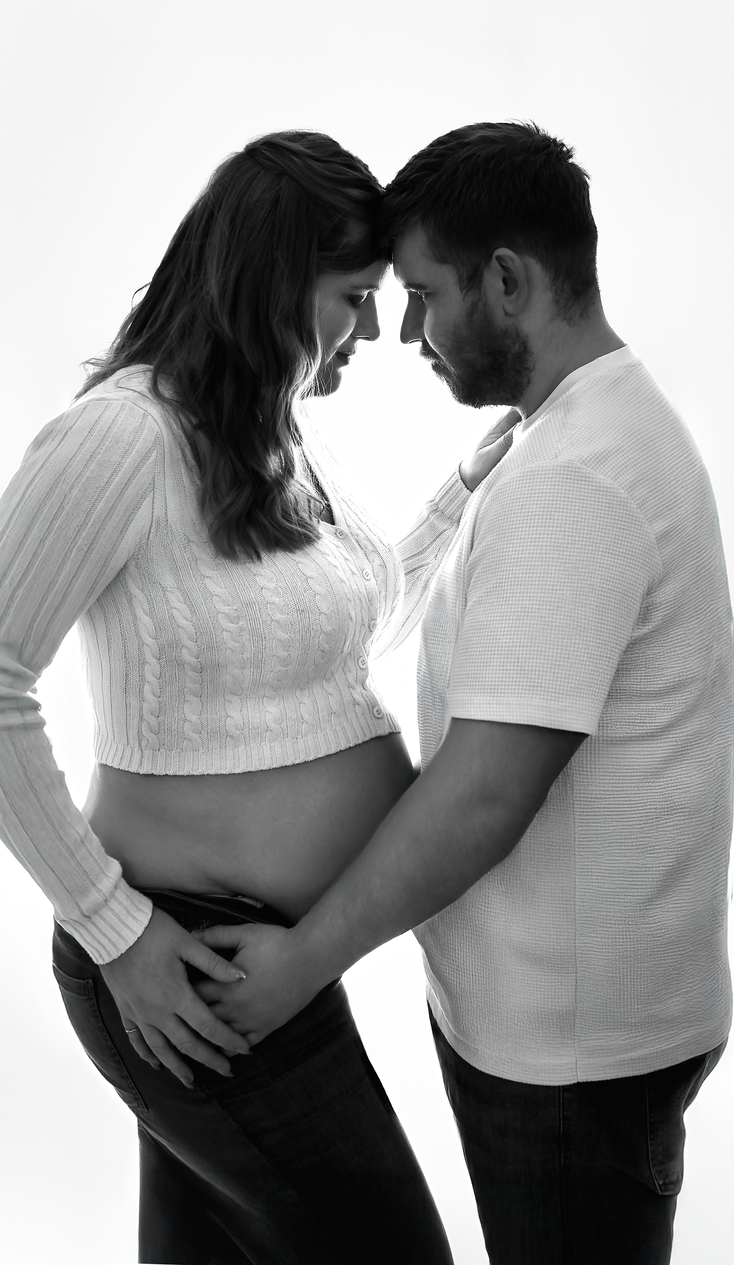 Black and white maternity photo of a couple touching foreheads, cradling a pregnant belly in a studio setting.
