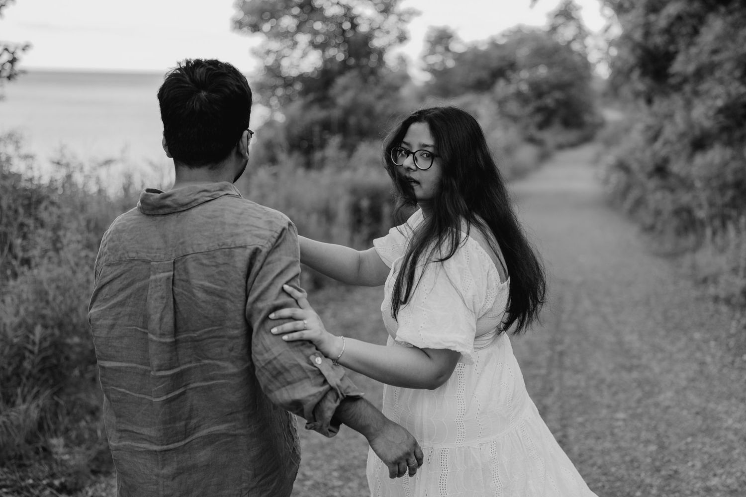 Black and white engagement photo of woman holding partner’s hand and looking back on trail