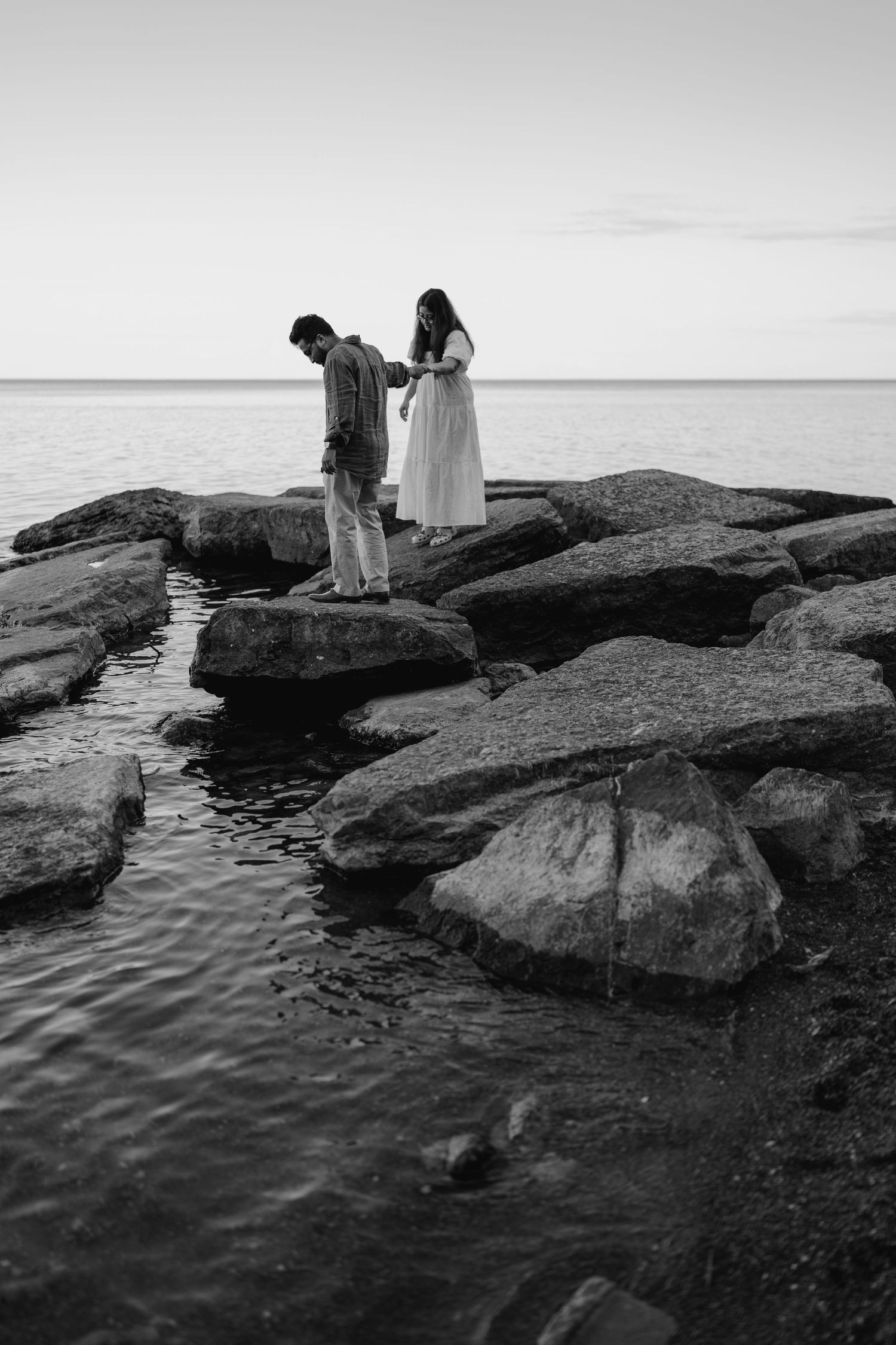 Black and white engagement photo of couple walking together on lake rocks