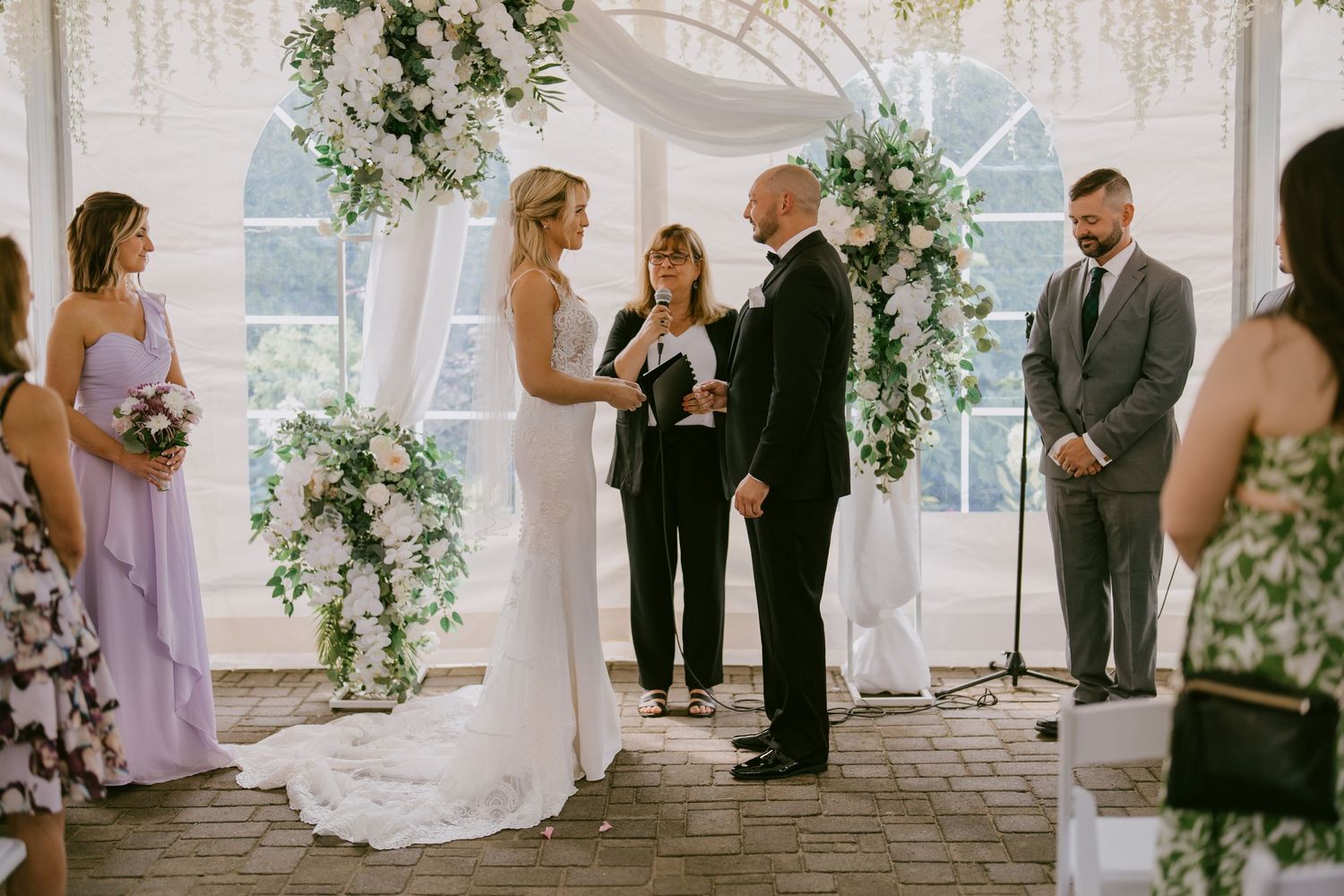 Bride and groom exchanging vows under a floral wedding arch at elegant tented ceremony  in Oshawa, available throughout Toronto and Durham r