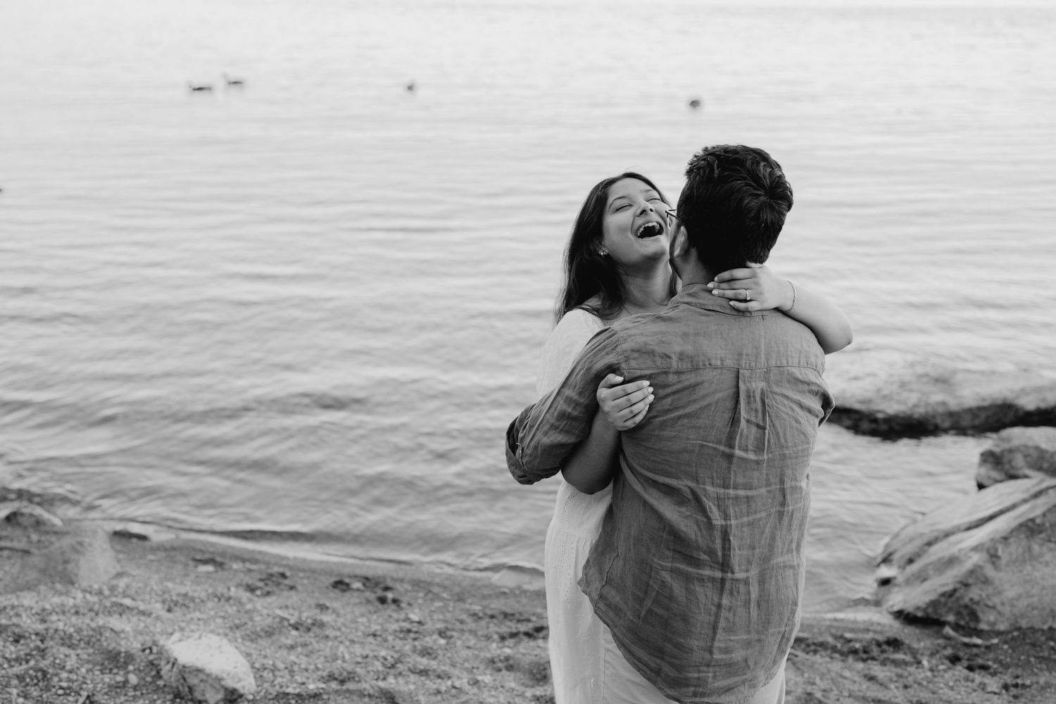 Candid black and white photo of woman laughing in partner’s arms by the water