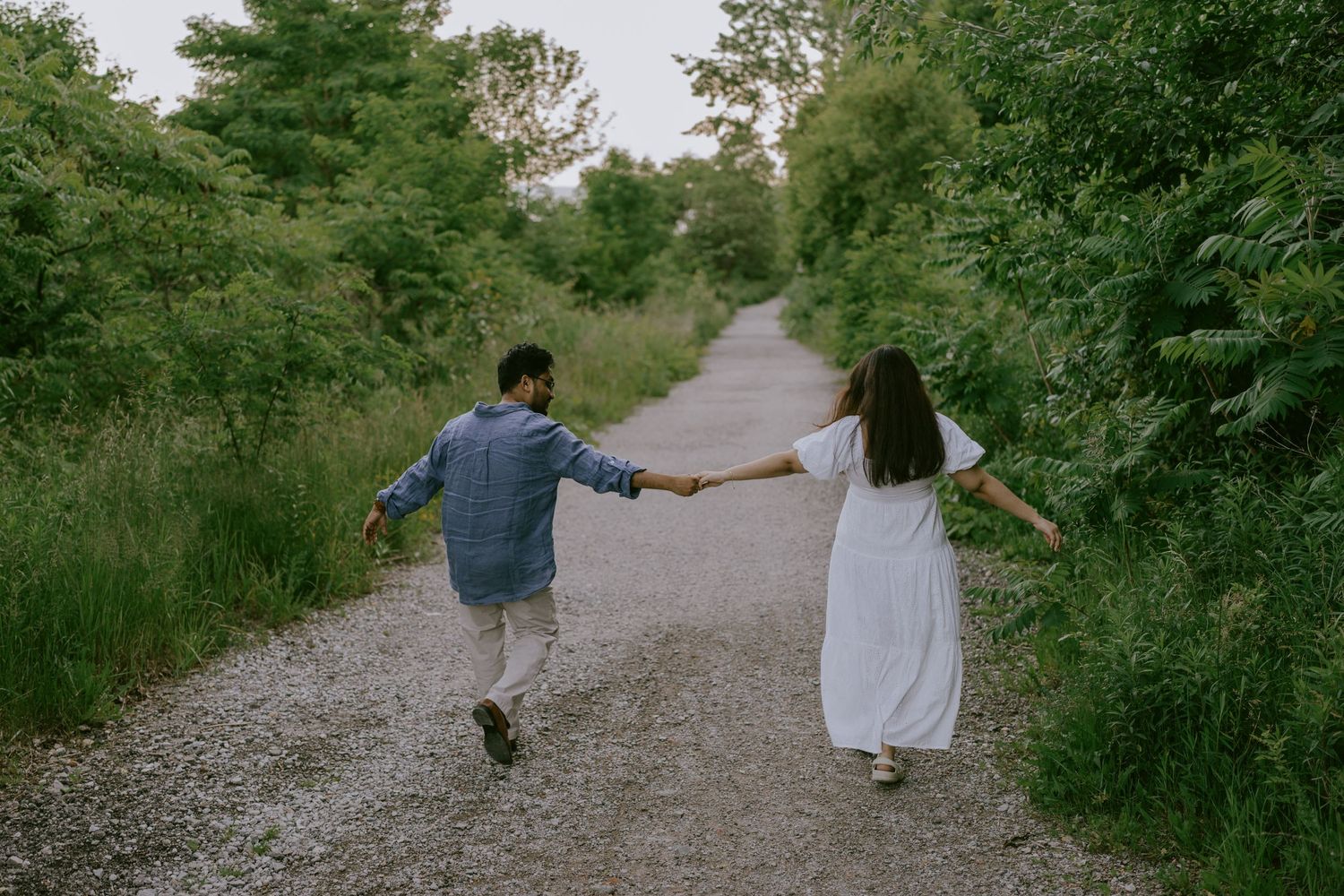 Engaged couple running hand-in-hand down gravel forest trail