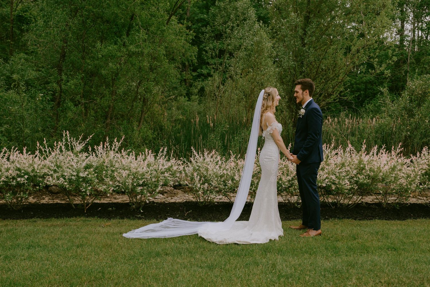 Bride and groom holding hands in garden with flowing veil on ground