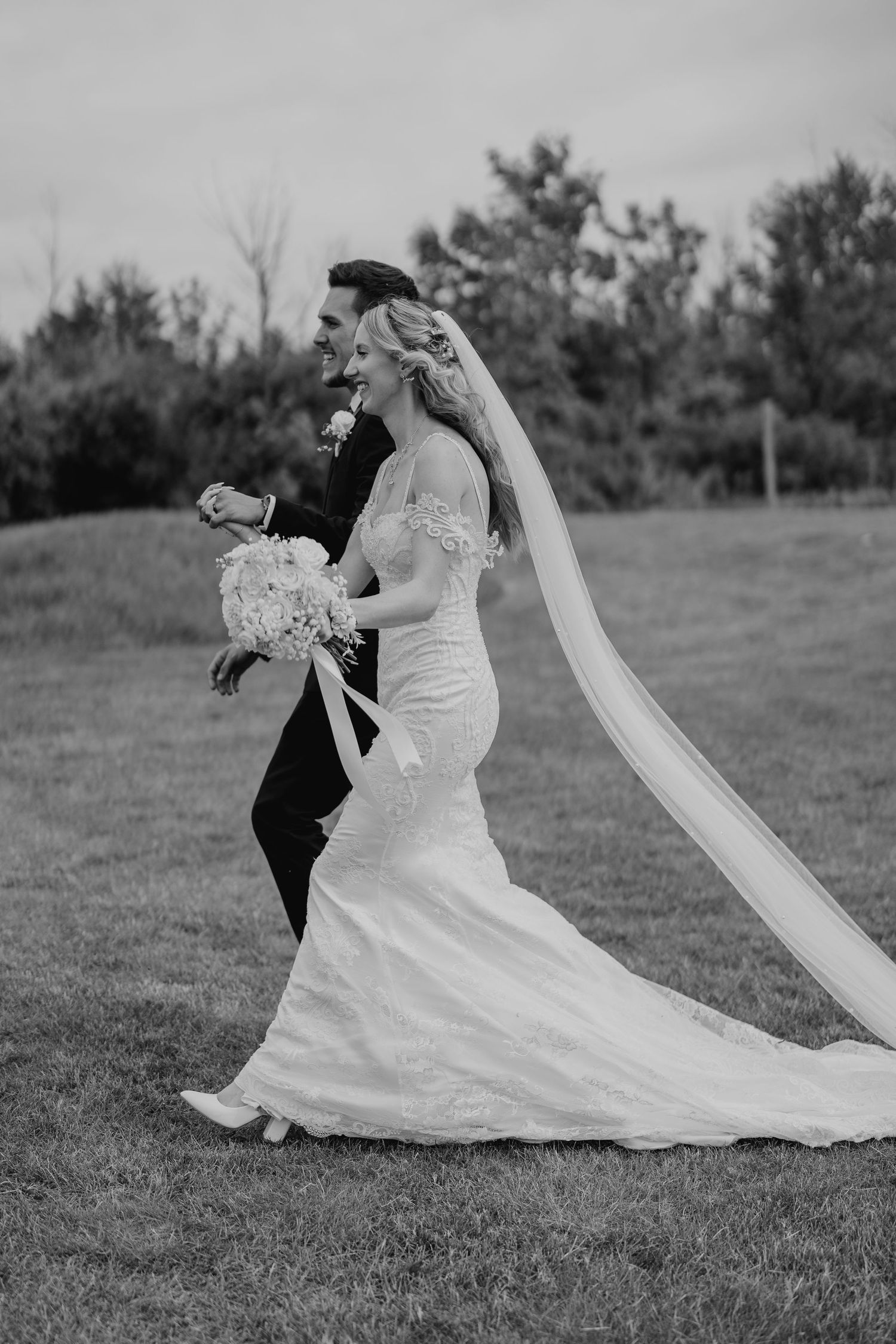 Black and white wedding photo of bride walking with flowing veil beside groom