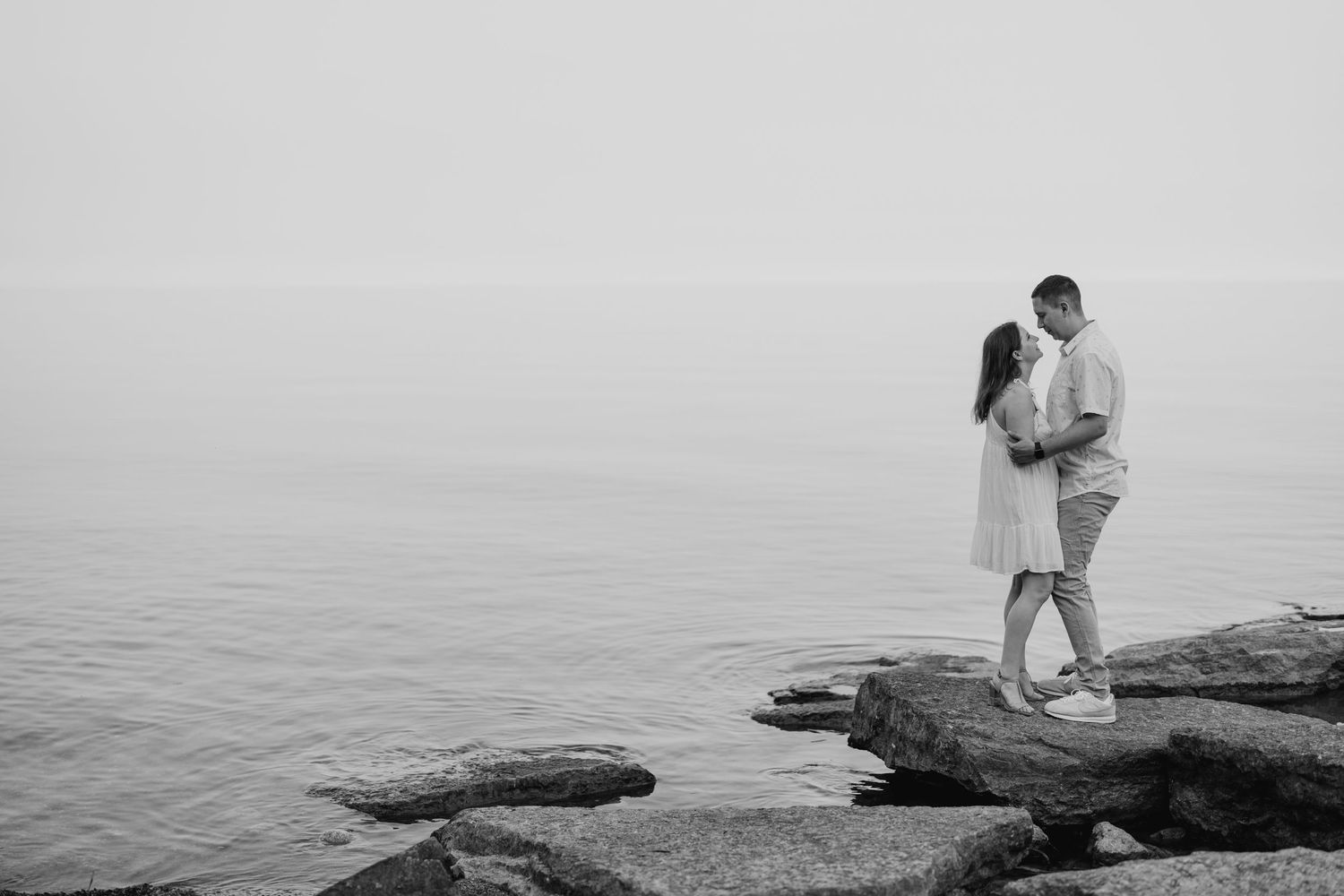 Black and white engagement photo of couple standing on lakeside rocks near lake Ontario in Oshawa, available throughout Toronto and Durham