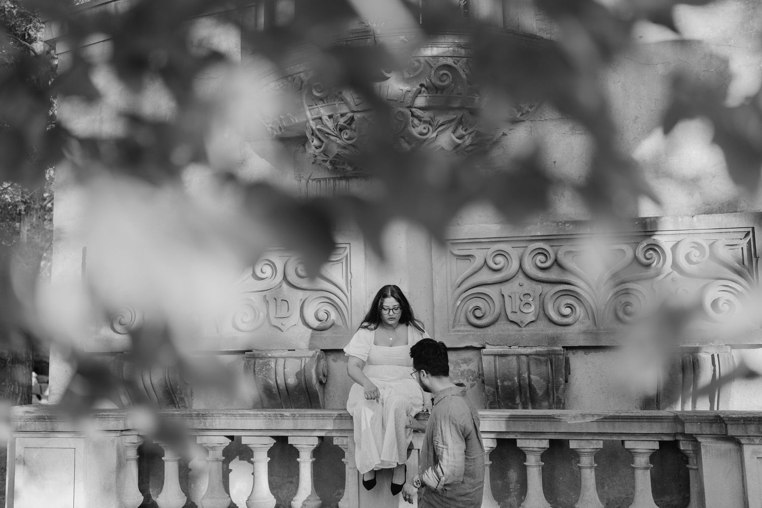 Black and white photo of couple walking behind ornate stone balcony with tree leaves in foreground