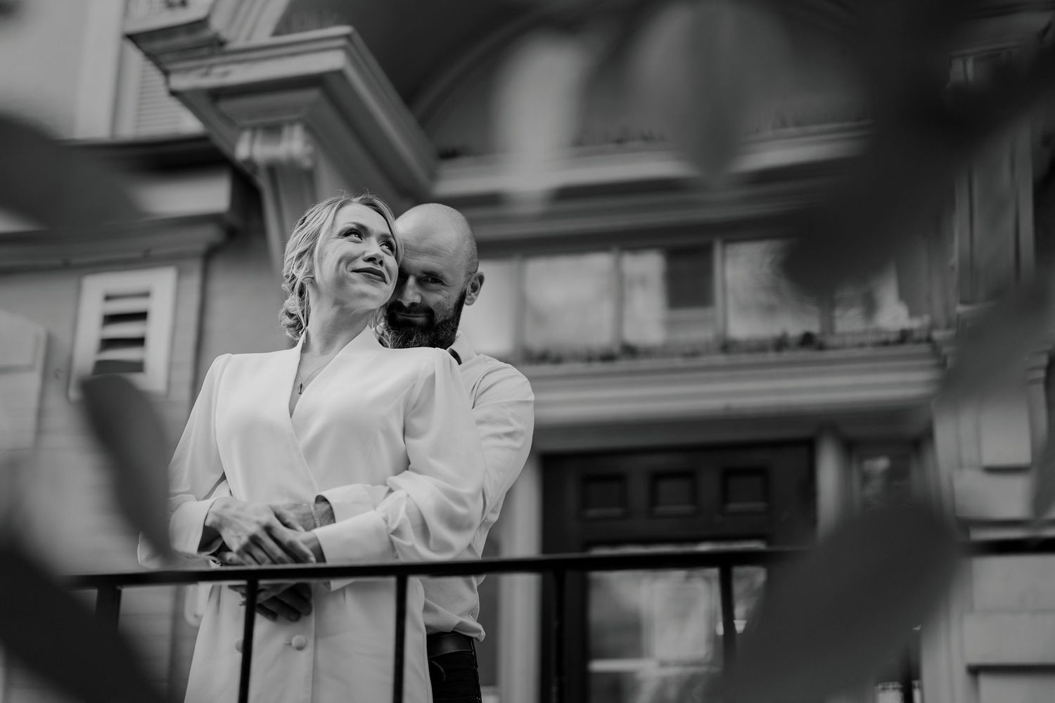 Black and white photo of couple embracing on balcony framed by soft leaves