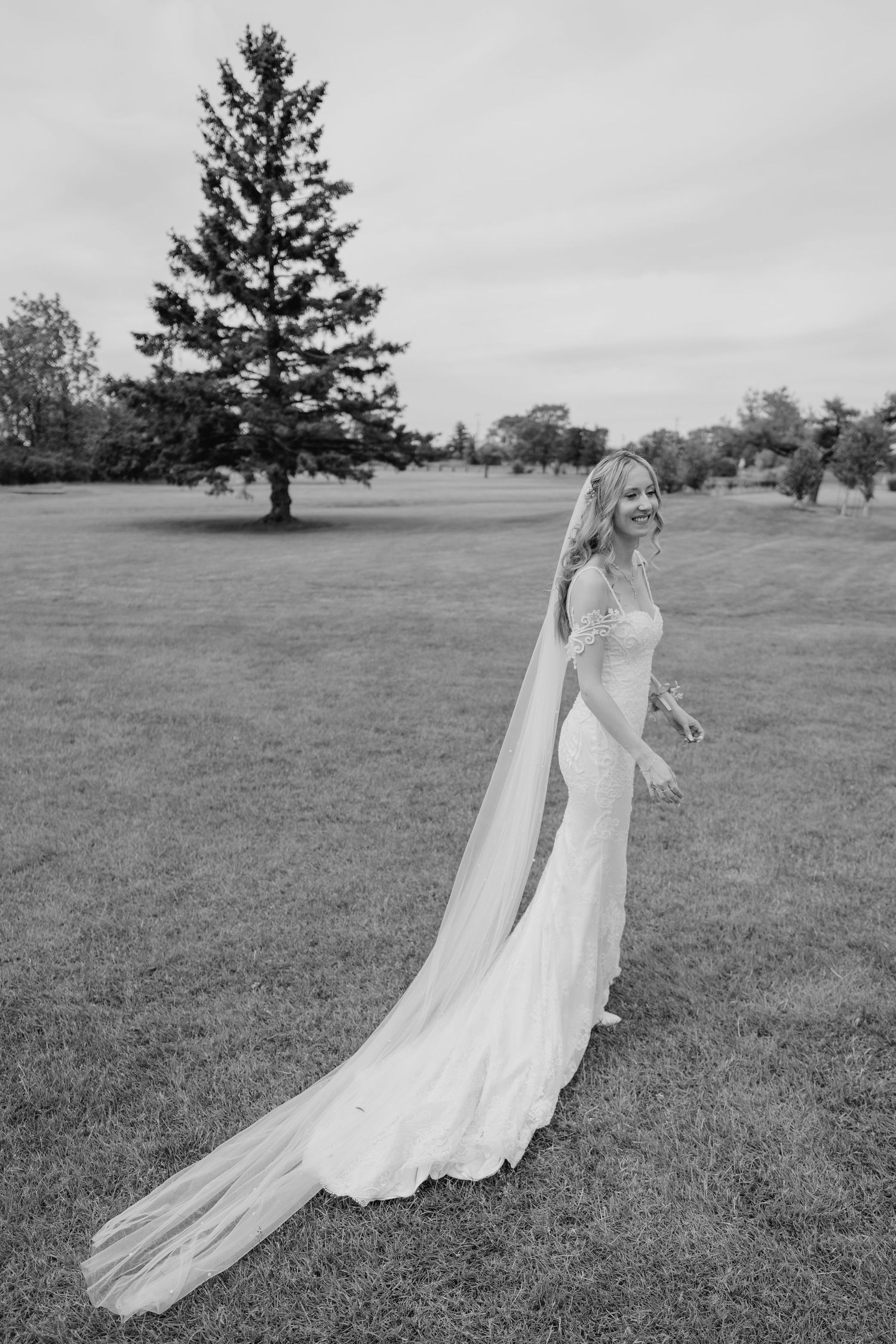 Black and white portrait of bride walking alone in open field with long train