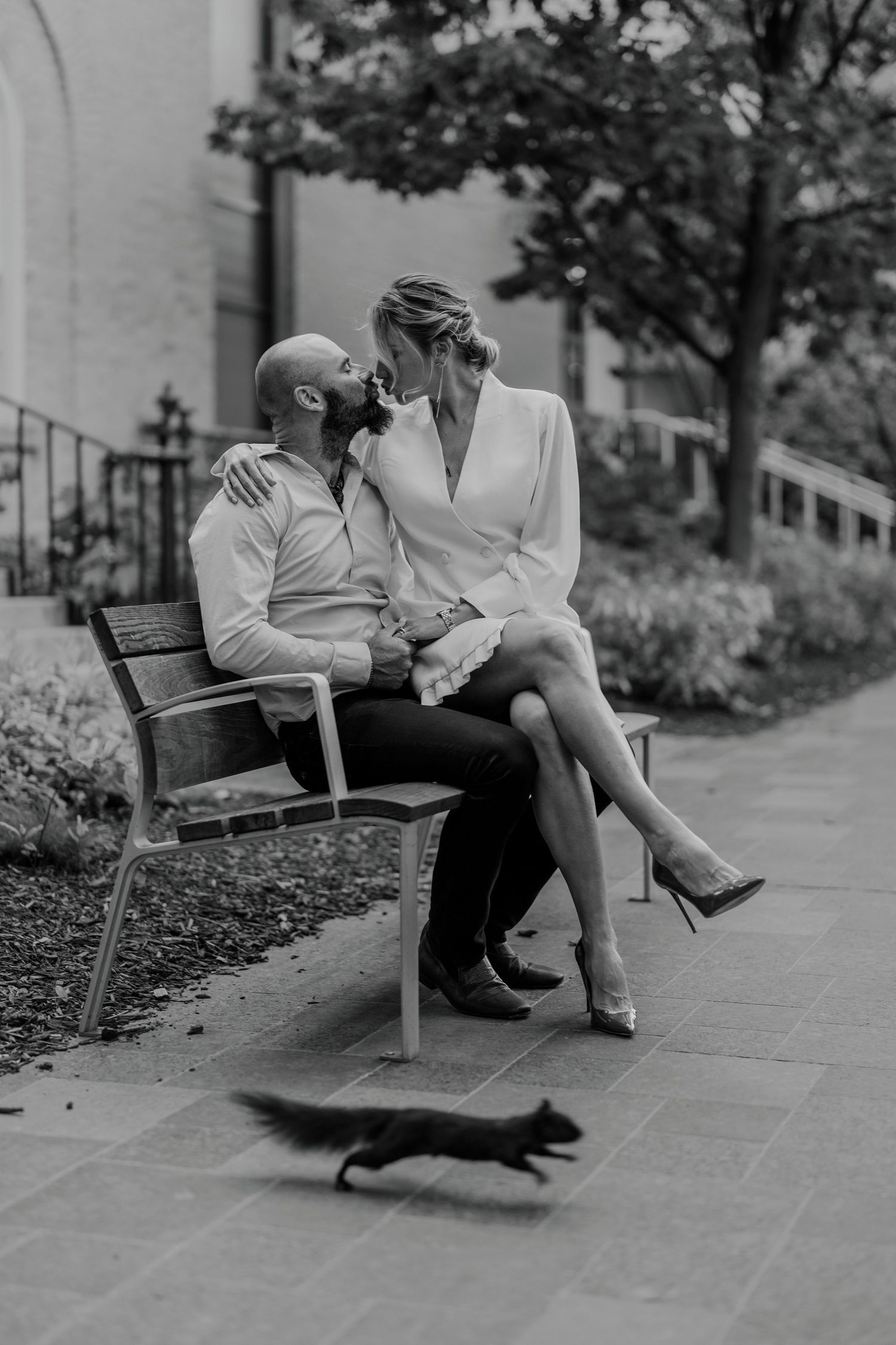 Candid black and white photo of couple sitting on park bench with squirrel passing by