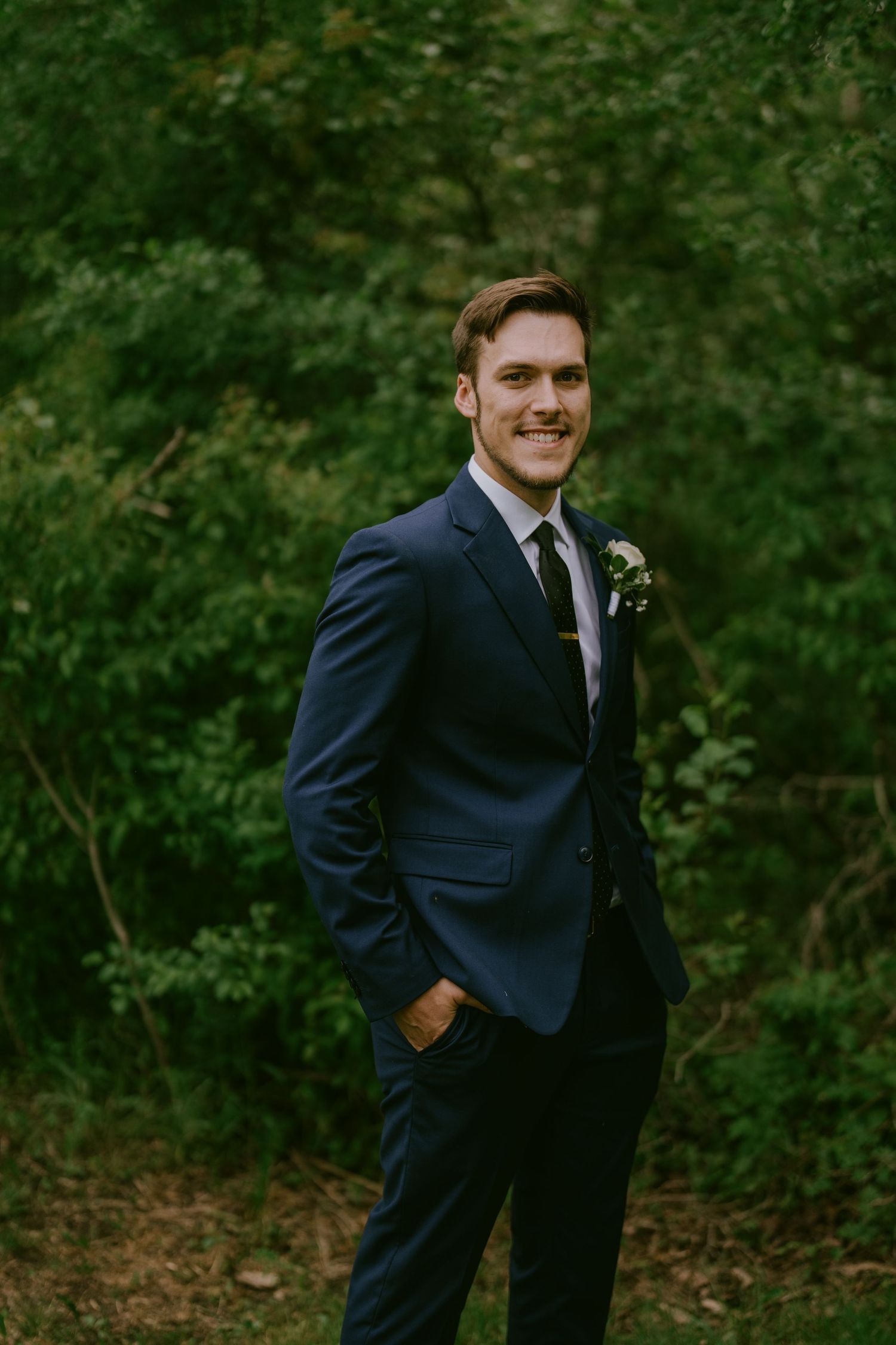 Groom standing alone in forested setting with hands in pockets