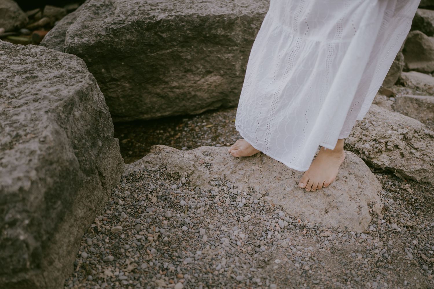 Close-up of bride’s bare feet and dress hem on lakeside rock
